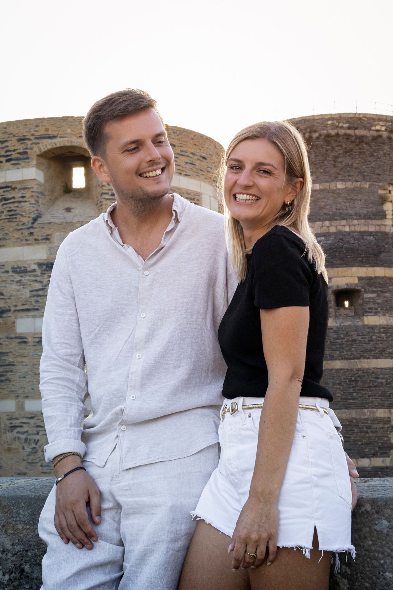 Couple souriant adossé à un mur de pierre, lui en chemise blanche, elle en haut noir, séance à Angers