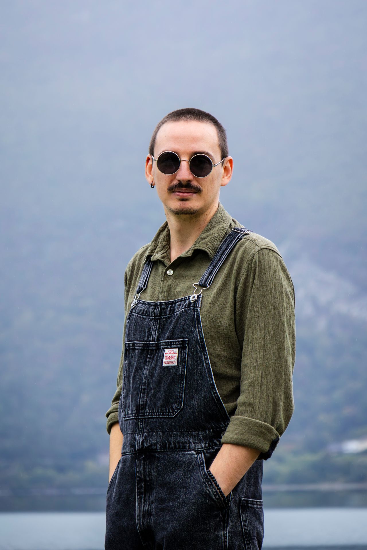 Portrait d'un homme en salopette, mains dans les poches et  lunettes de soleil, regardant l'objectif avec lac et montagne, séance en Savoie