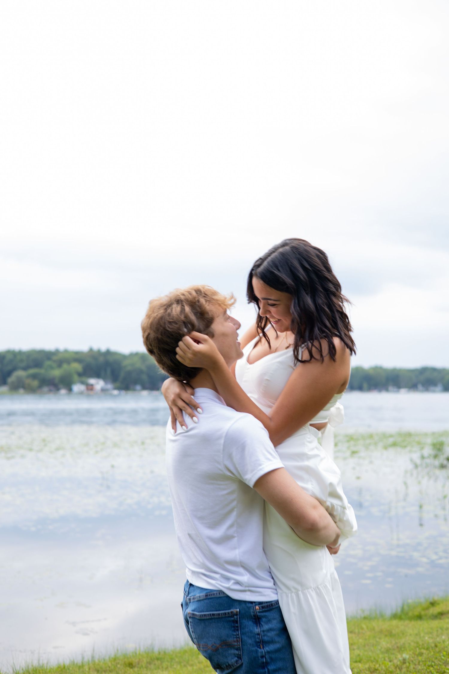 Romantic couple embracing by a lakeside, man lifting woman in white dress, sharing a tender moment outdoors.