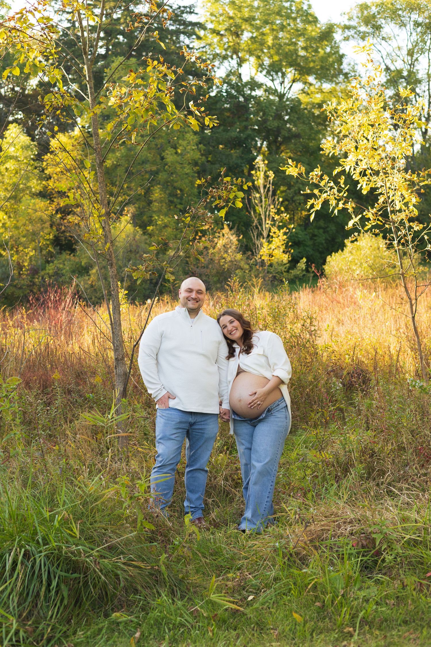 Couple in white sweaters and jeans posing outdoors in autumn field, pregnant woman cradling baby bump, leaning on partner.