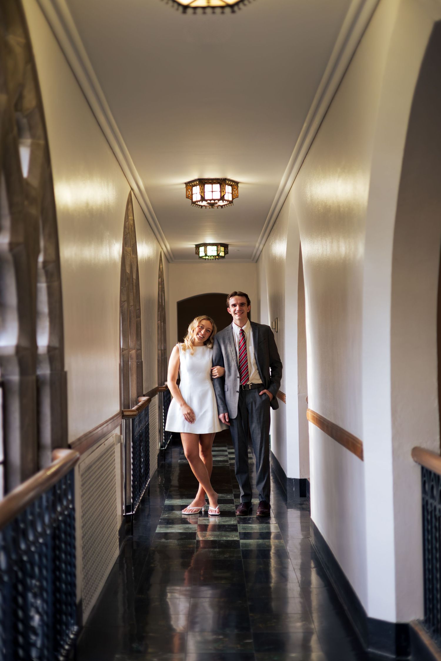 Elegant couple walking hand in hand through a grand hotel corridor with ornate ceiling lights and dark marble floors.