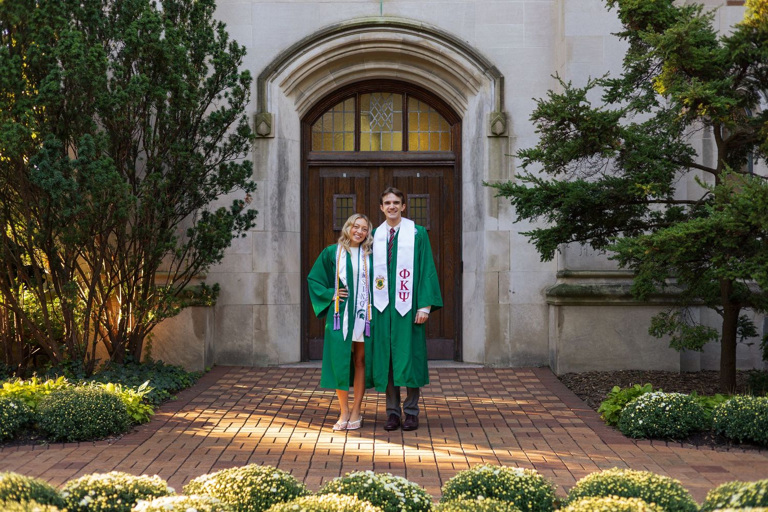 Two graduates in green robes pose proudly before an arched stone building entrance on a brick pathway.