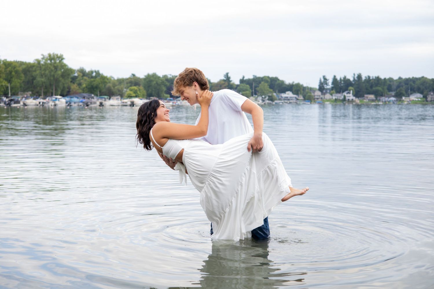 Couple in white attire sharing a romantic moment standing in a lake with trees and boats in the background.