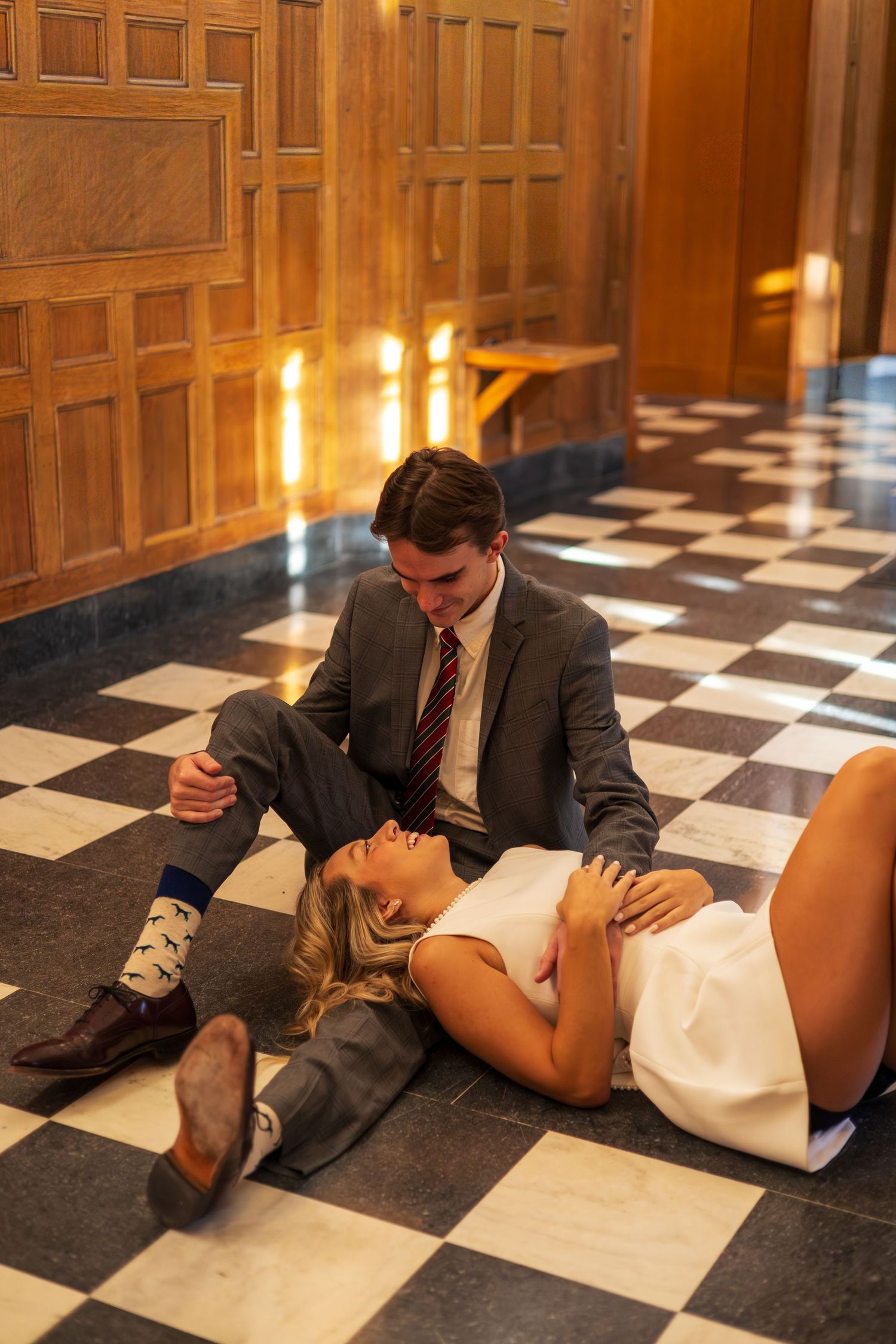 Couple in formal attire posing on checkered marble floor in elegant wood-paneled hall.