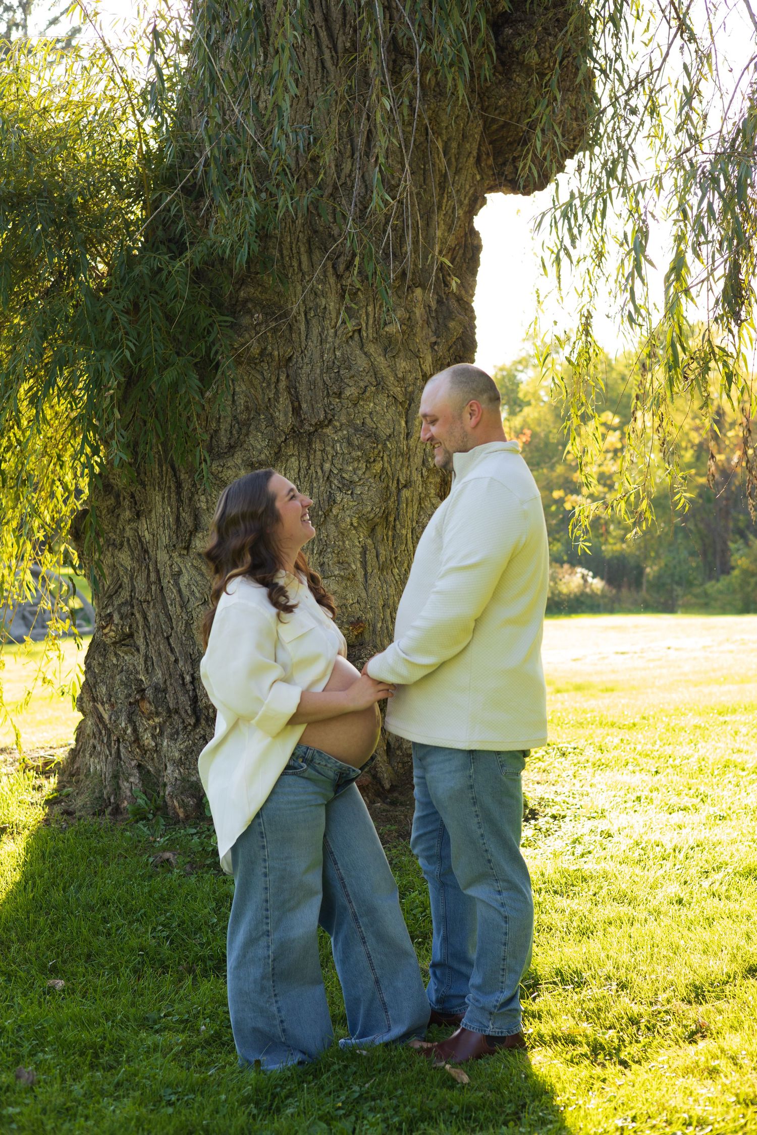 Pregnant couple holding hands and smiling at each other outdoors near a large willow tree on a sunny day.