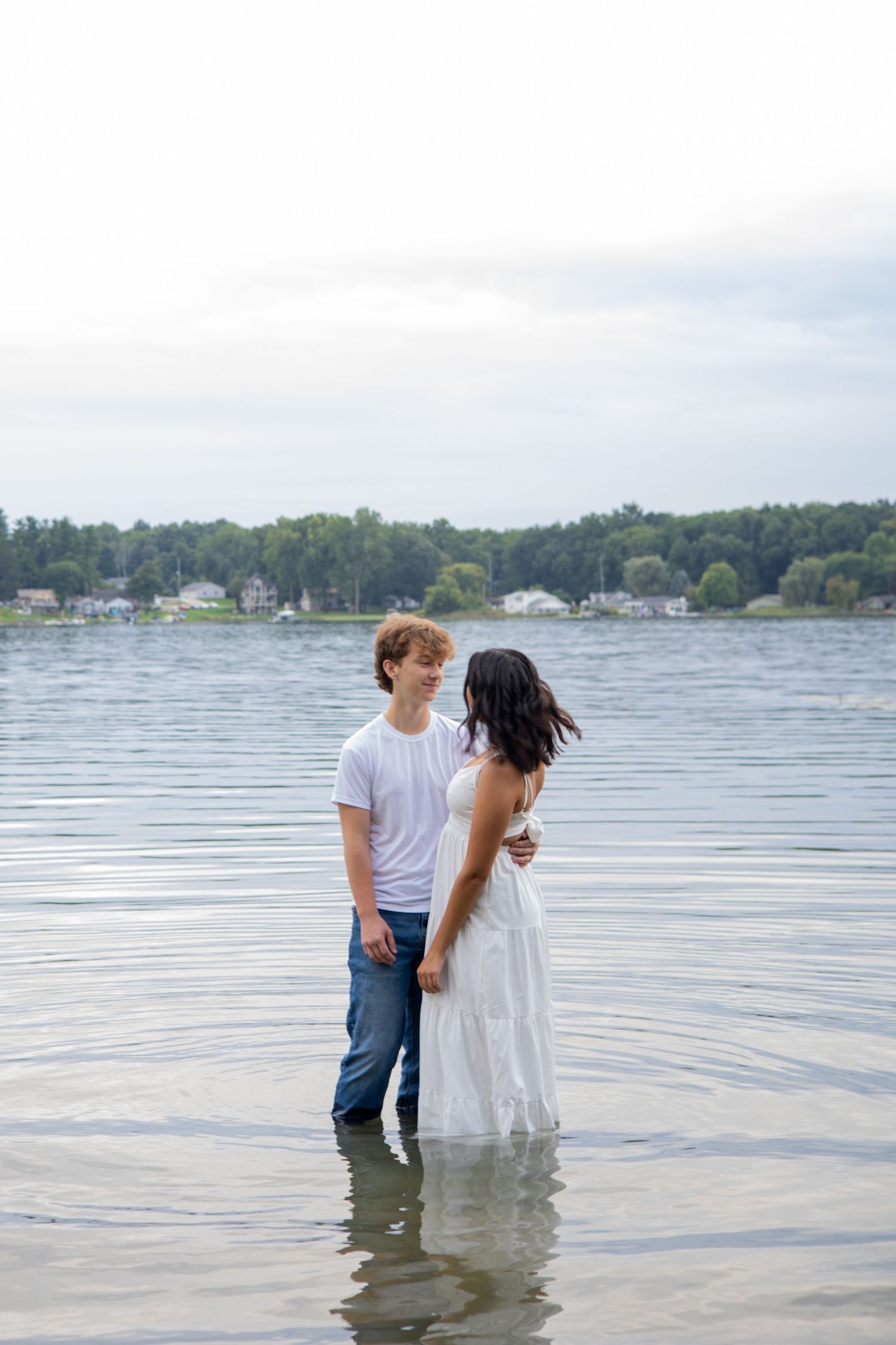 A young couple stands romantically in a lake, the man in a white tee and jeans, the woman in a flowing white dress.