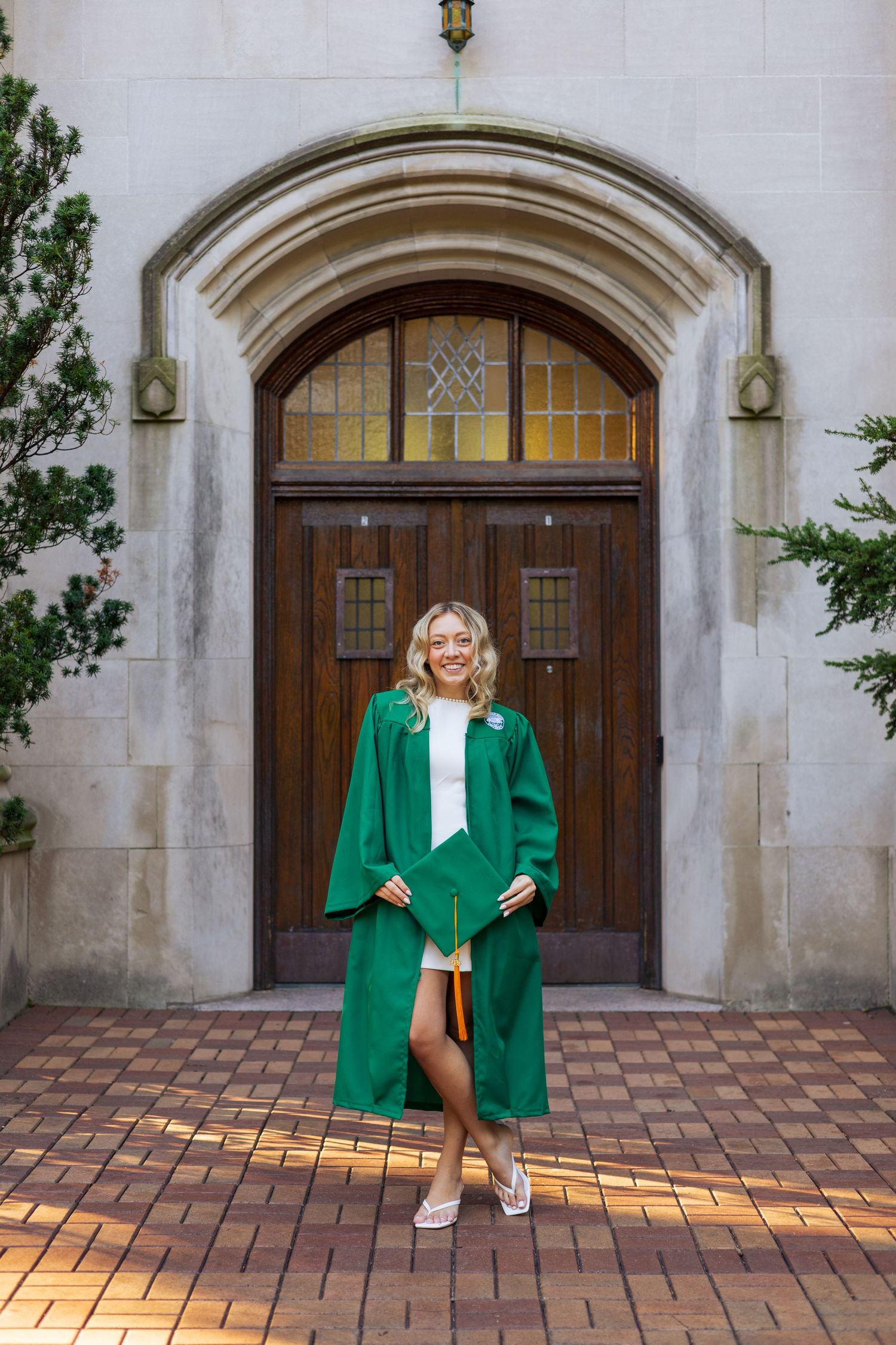 Smiling graduate in green cap and gown posing in front of an arched wooden door on a brick pathway.