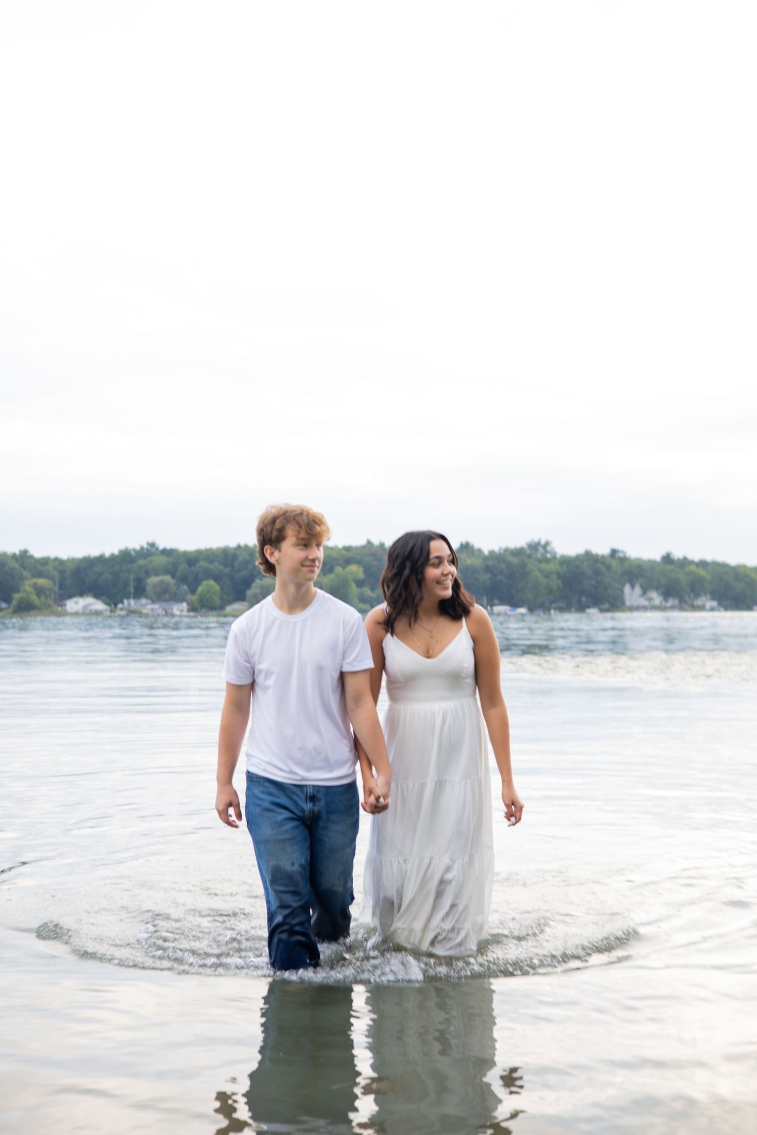 Couple holding hands wading through shallow lake water, woman in white dress and man in jeans and white tee.