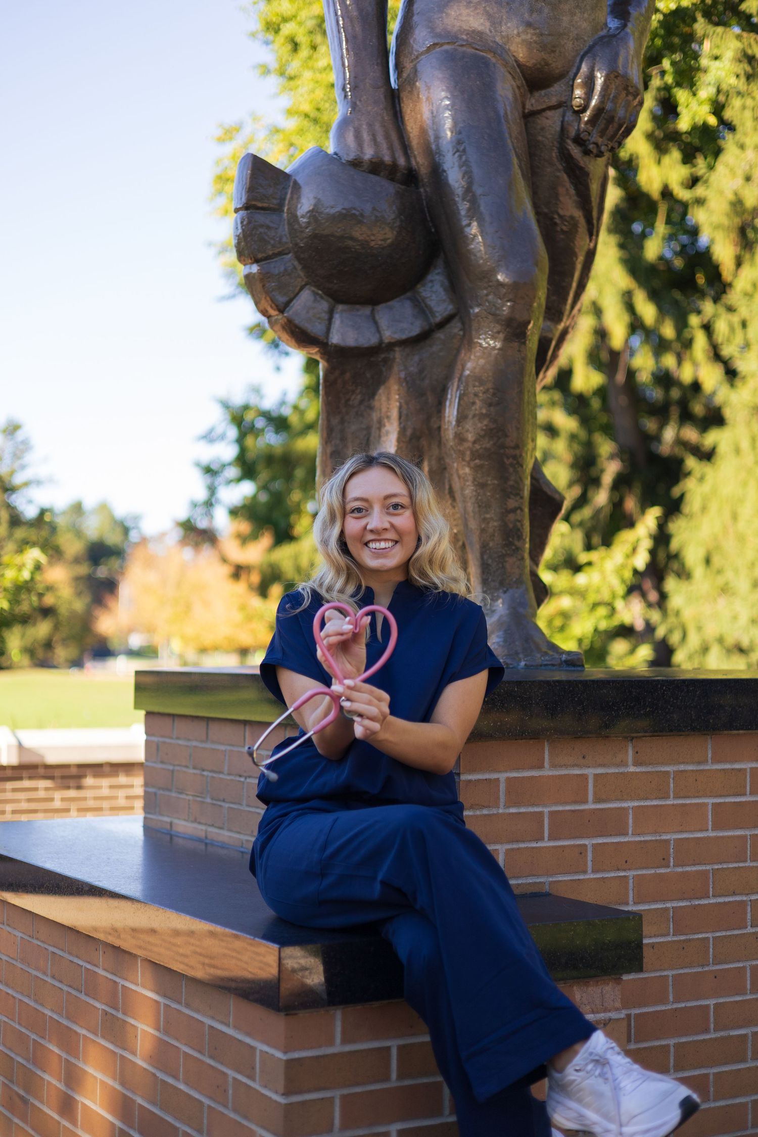 Smiling nursing student in blue scrubs holds a pink stethoscope heart shape while sitting by a bronze statue outdoors.