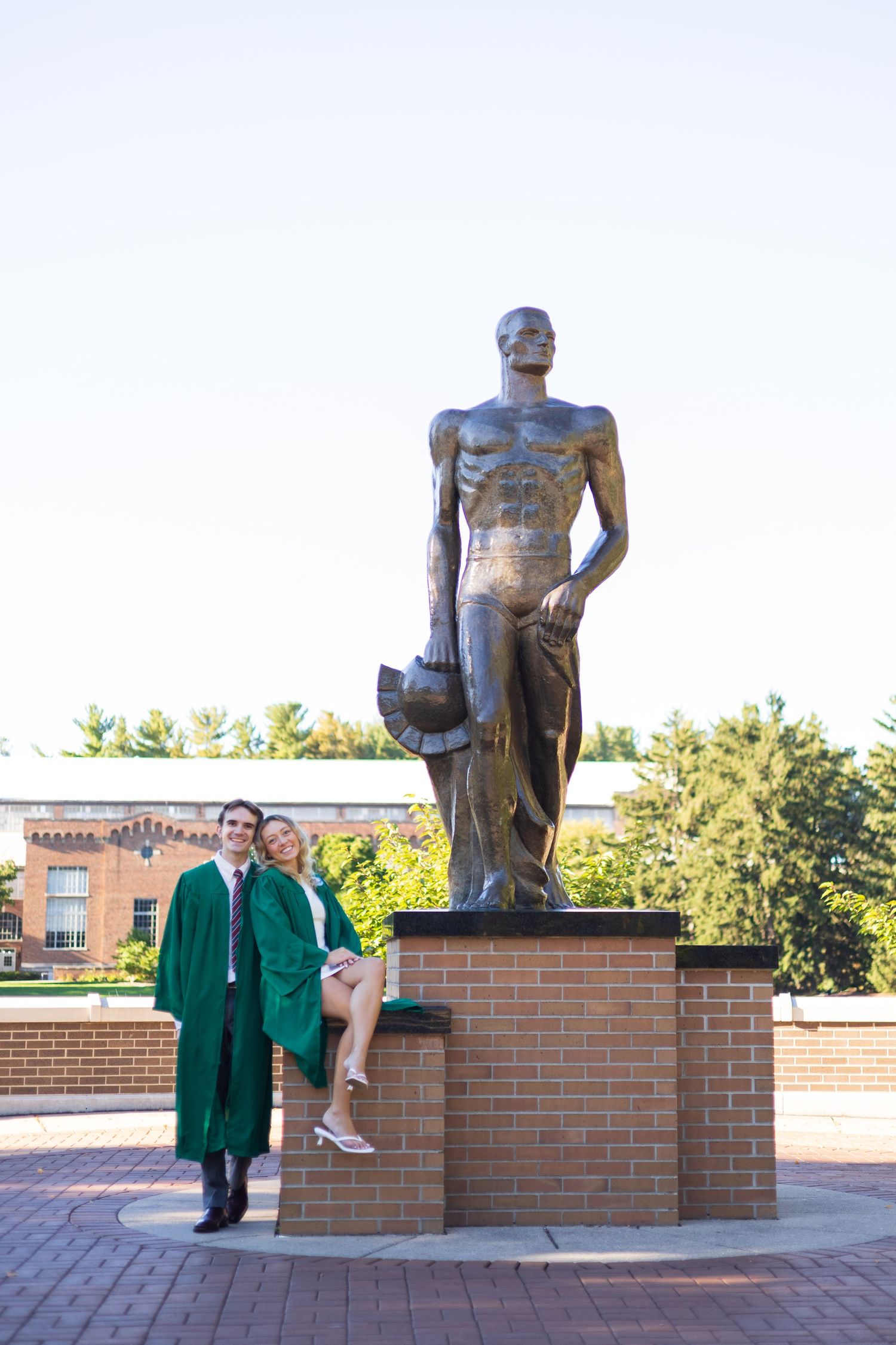 Graduate in green gown poses with companion beside the Spartan statue at Michigan State University campus.