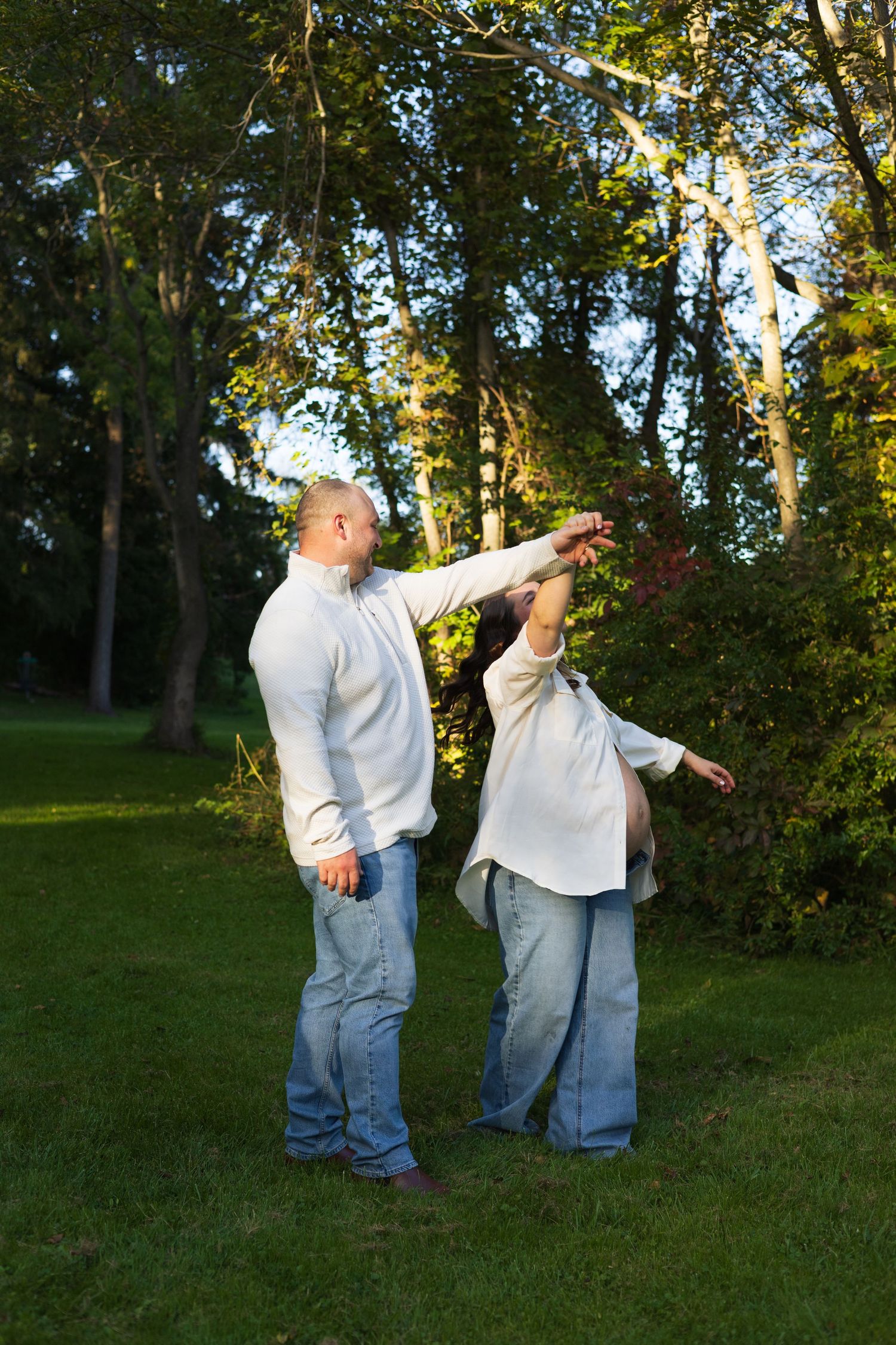 A man and woman in matching white shirts and jeans dance together on green grass in a sunlit park.