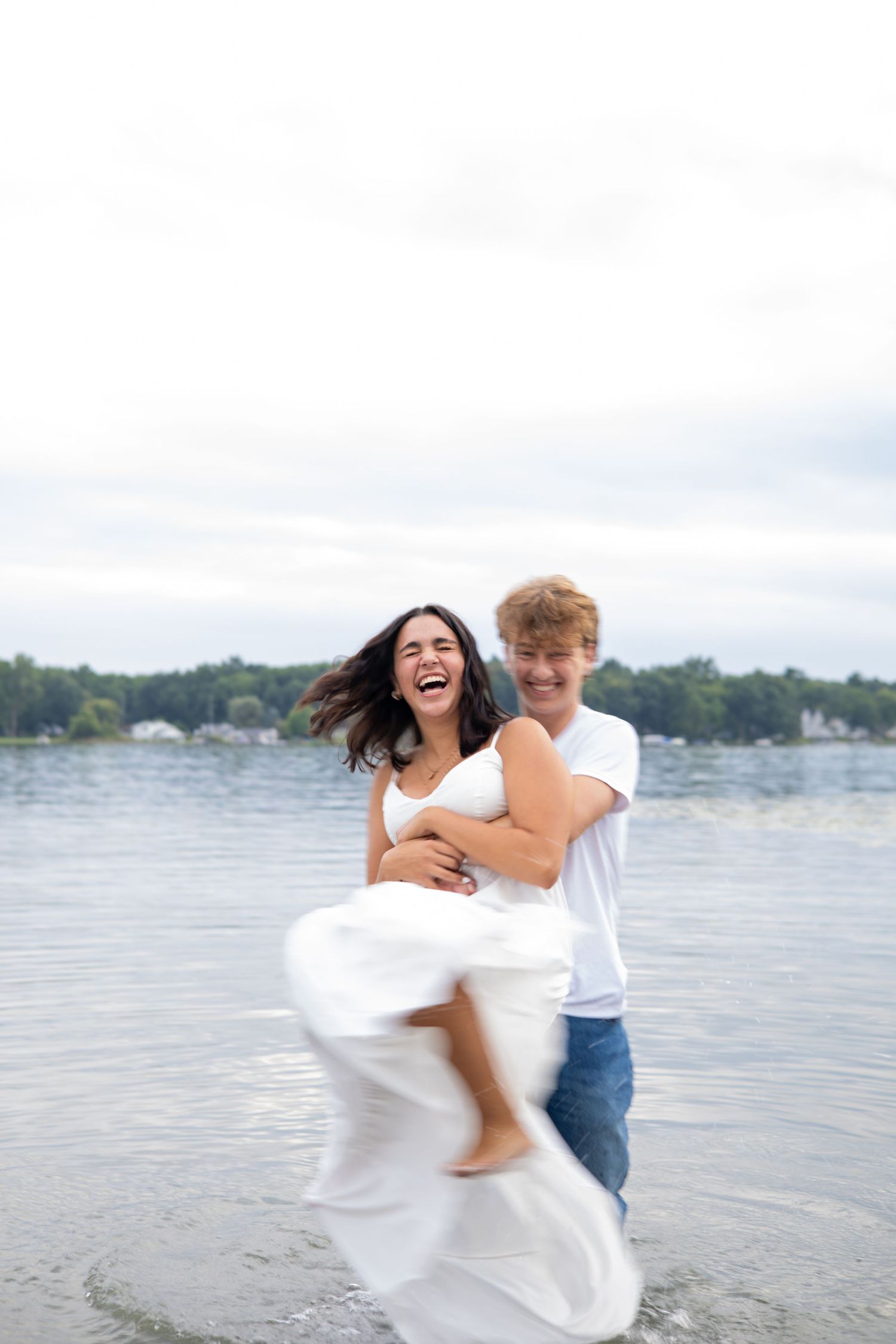 A joyful couple laughs and spins together on a lakeside beach, the woman in a white dress lifted off the ground.