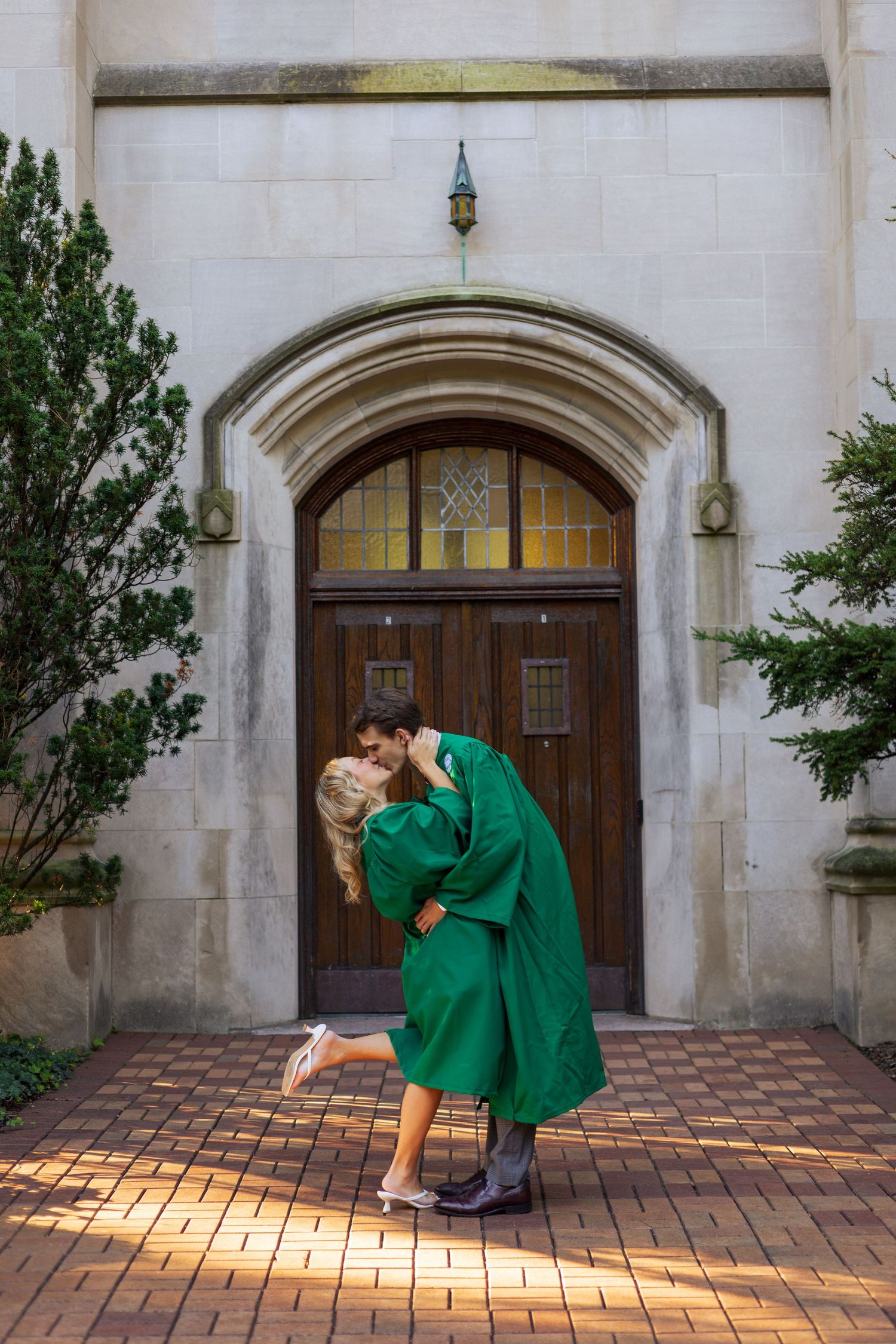 Couple sharing a romantic dip kiss in front of ornate stone archway, woman wearing green graduation gown.