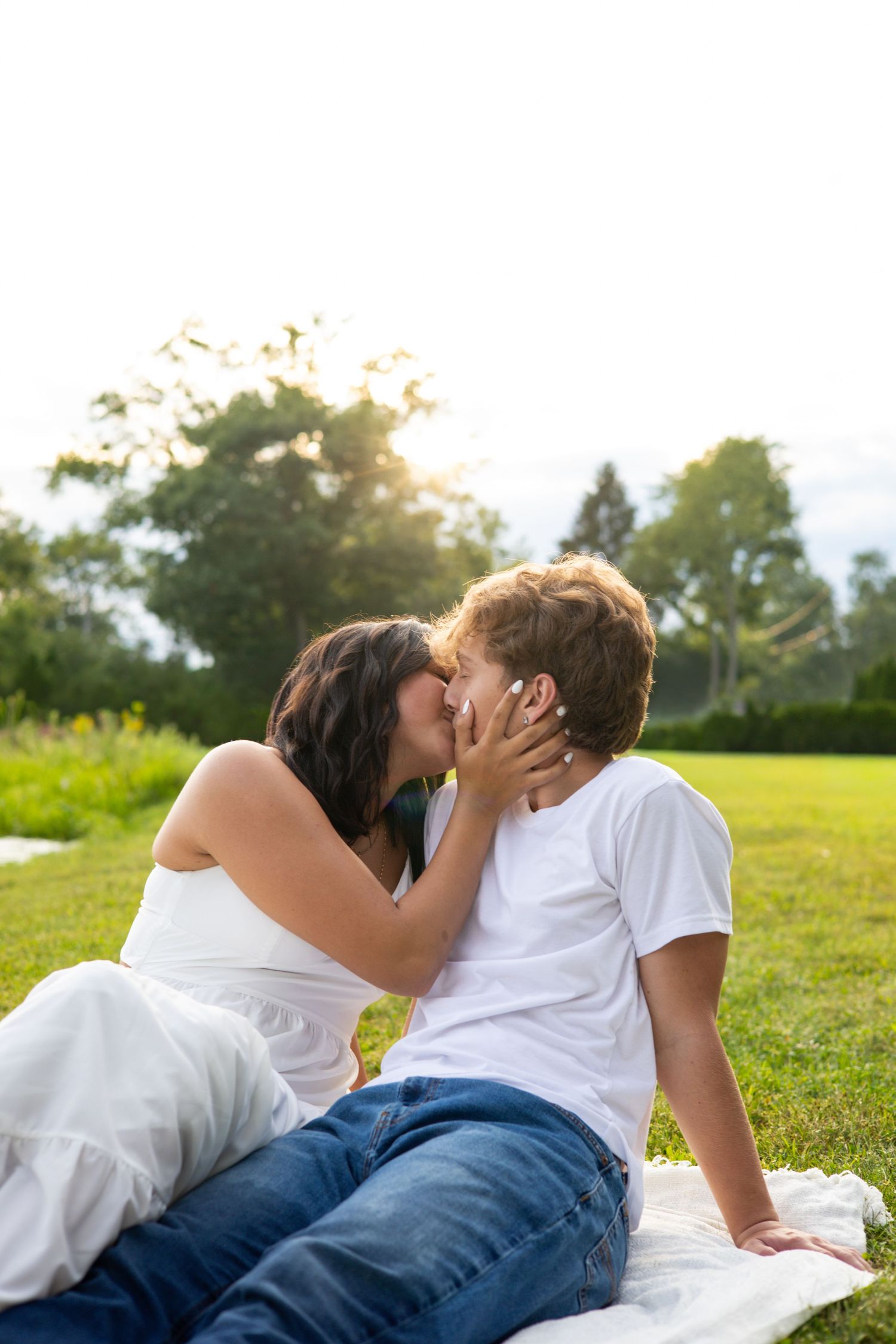 Romantic couple kissing on a blanket in a sunny green park, woman in white dress and man in white t-shirt and jeans.