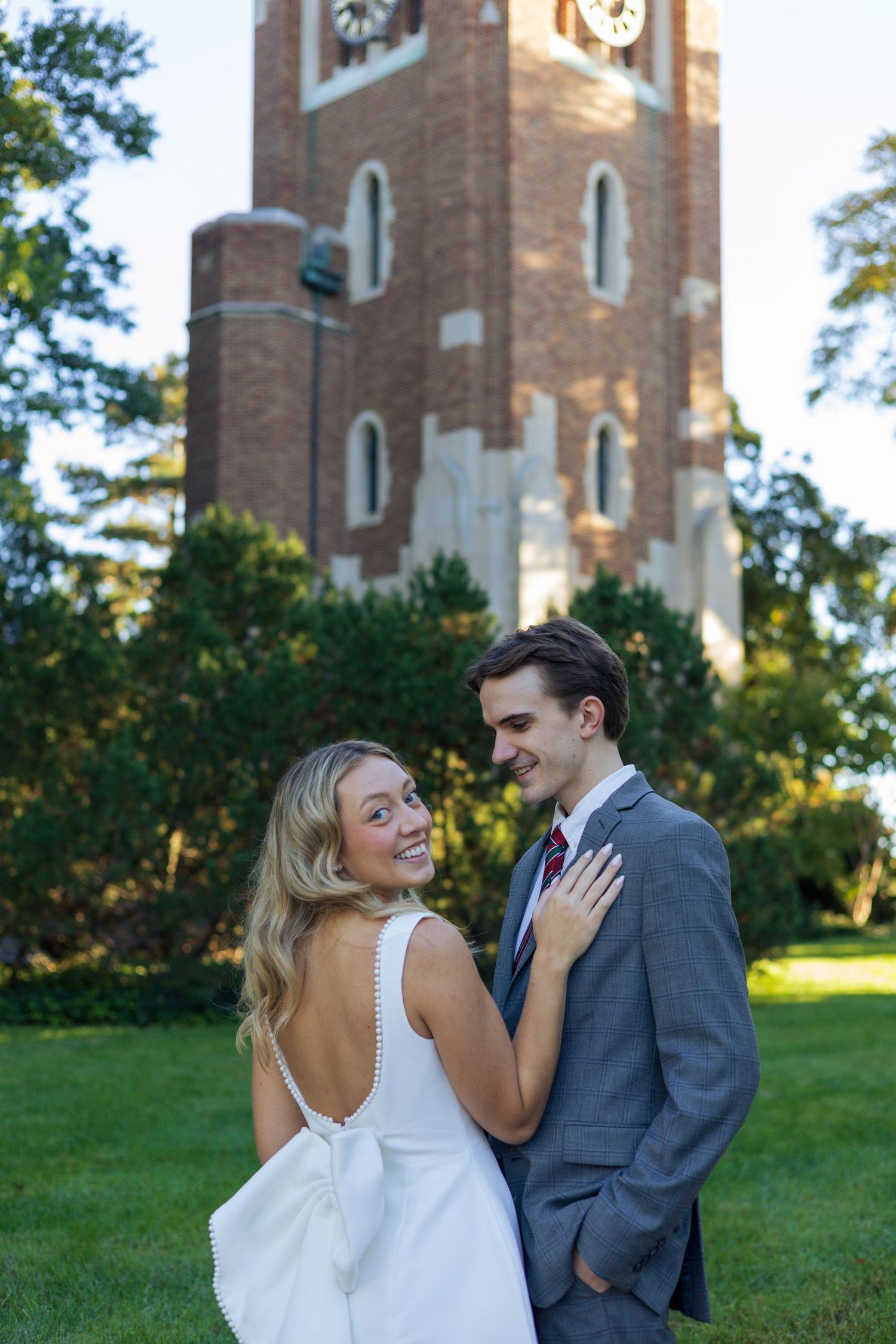 A smiling couple in formal attire poses outdoors near a brick clock tower on a green lawn.