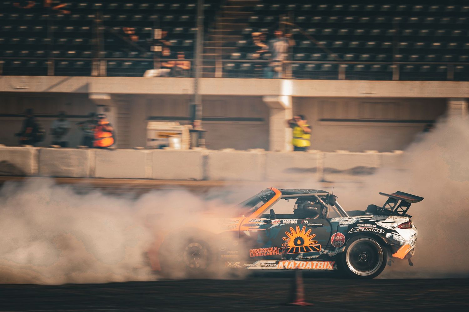 Orange and black drift car engulfed in tire smoke while performing a drift at a stadium event.