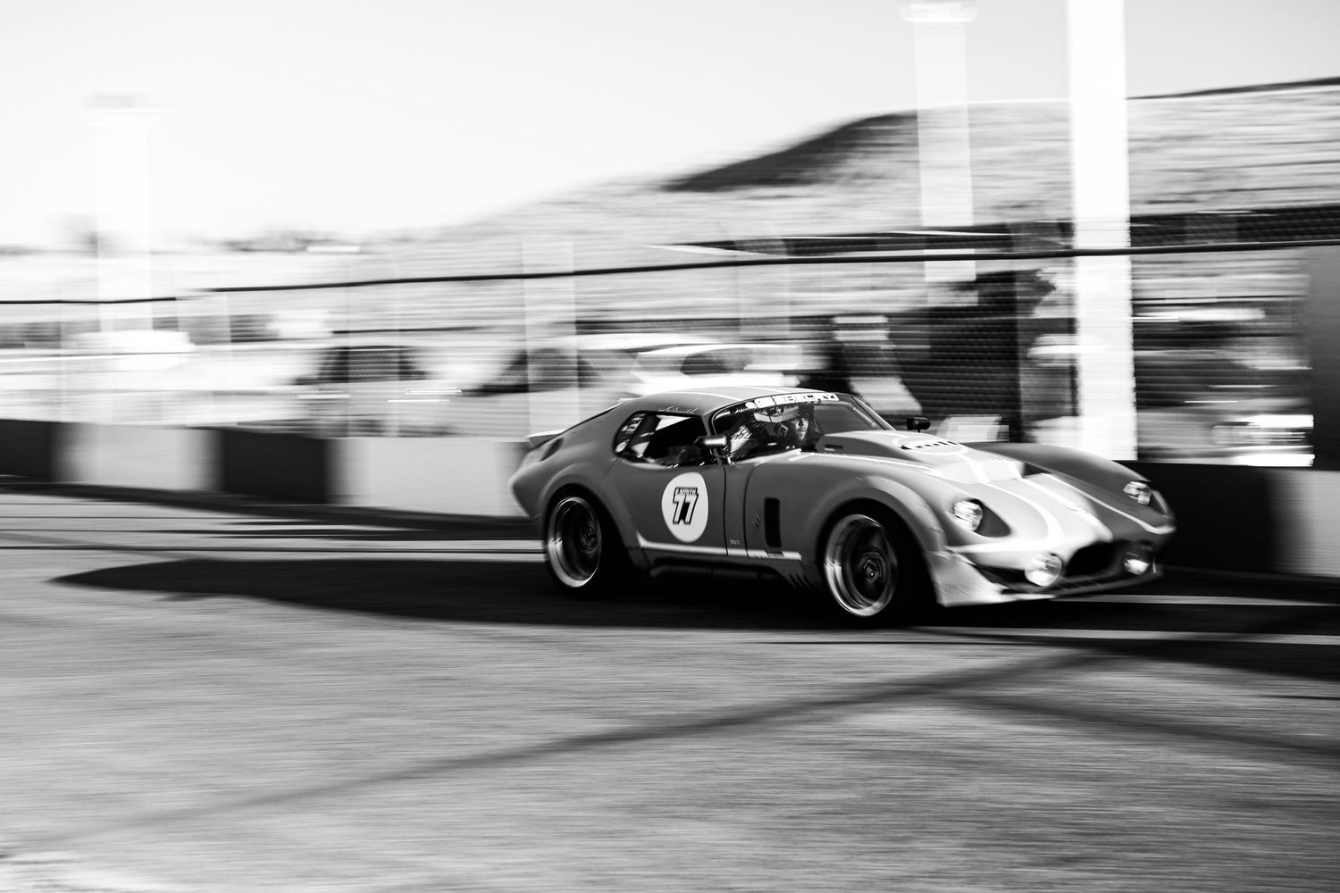 Black and white panning shot of a vintage sports car racing past pit lane barriers.