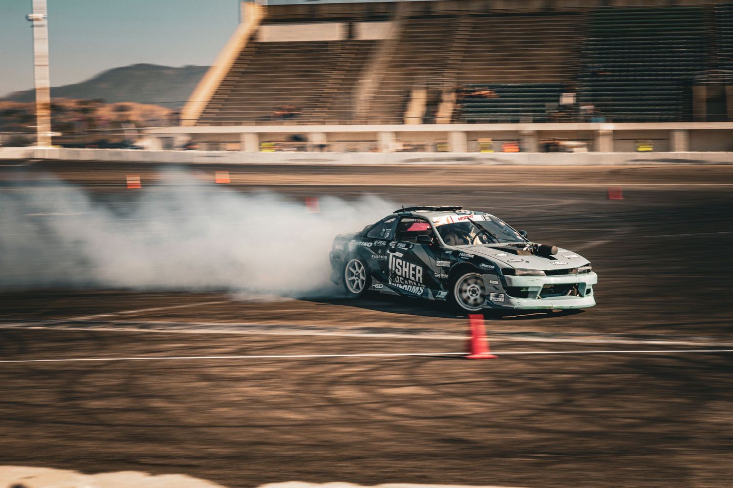 Black drift car producing thick smoke while navigating around an orange cone on a track.
