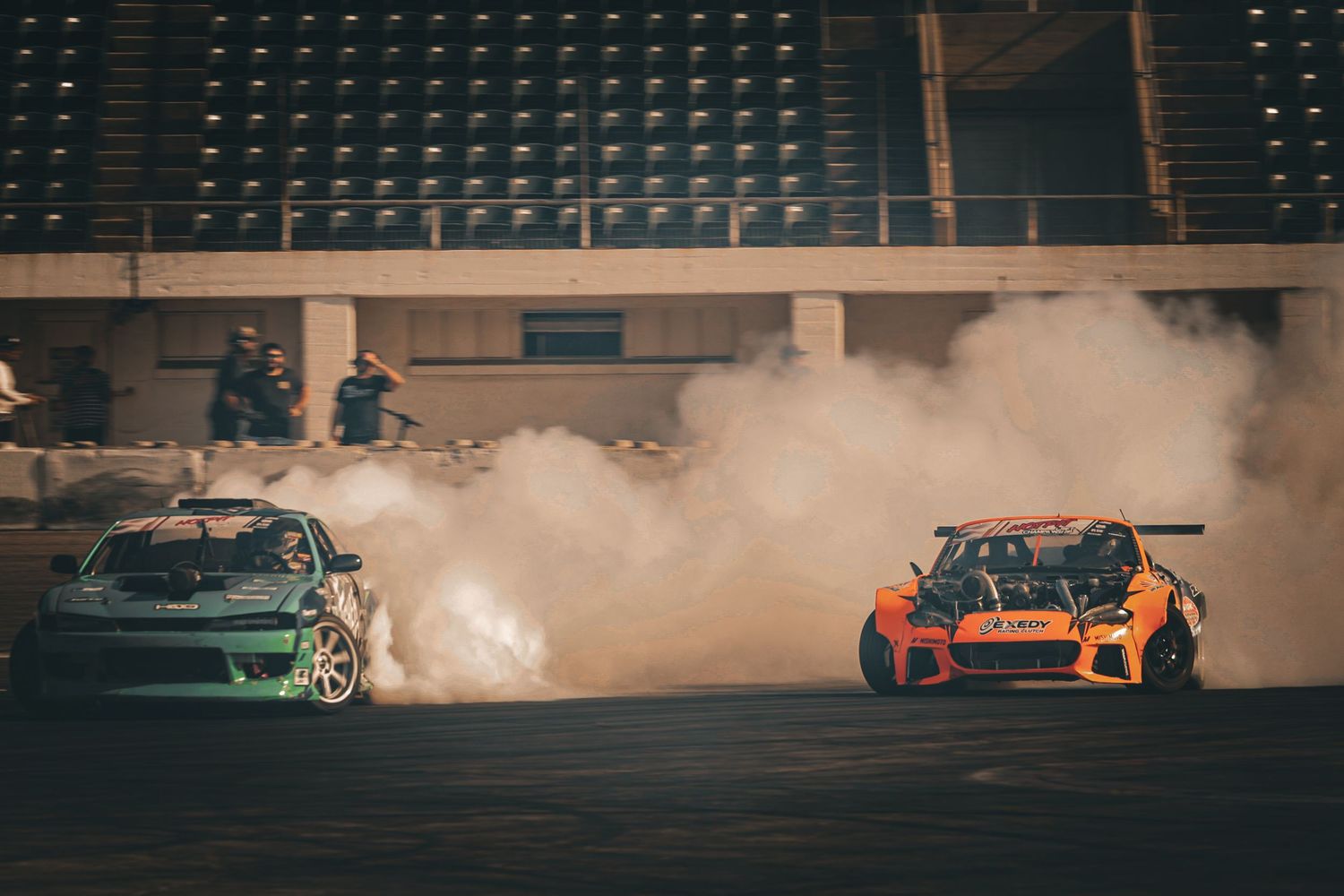 Two drift cars surrounded by thick smoke compete on a race track with empty grandstands in the background.