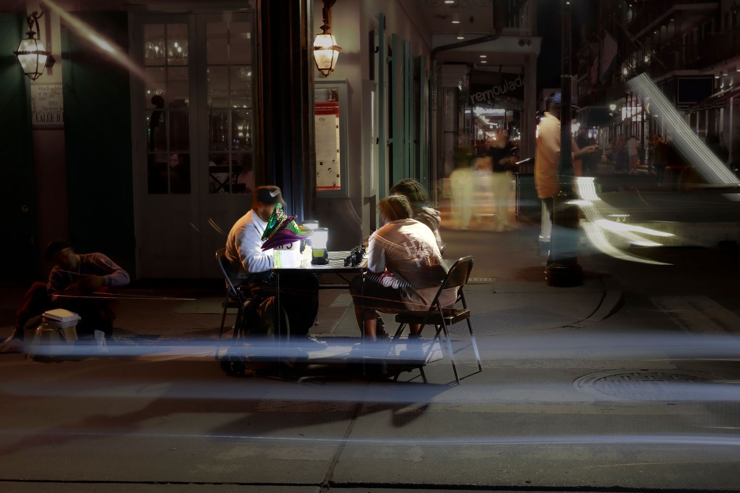 Two people dine at an outdoor cafe table at night, with a blurred figure passing by.