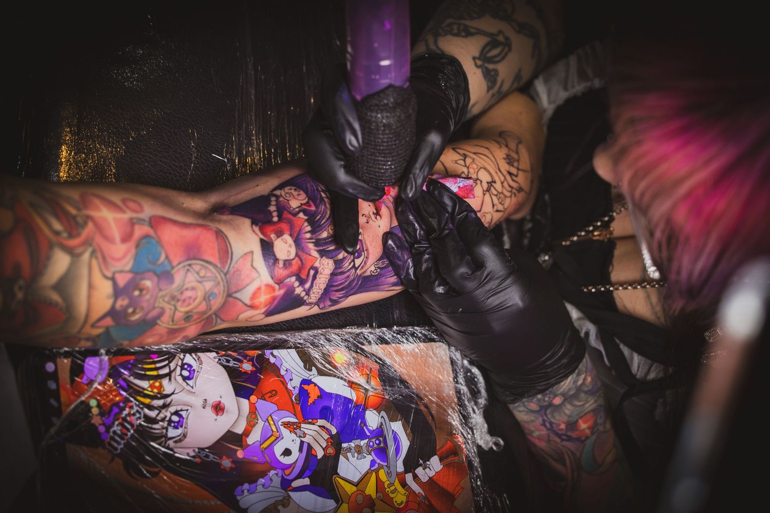 Tattoo artist applying colorful anime-style tattoo on a heavily tattooed arm in dark studio.