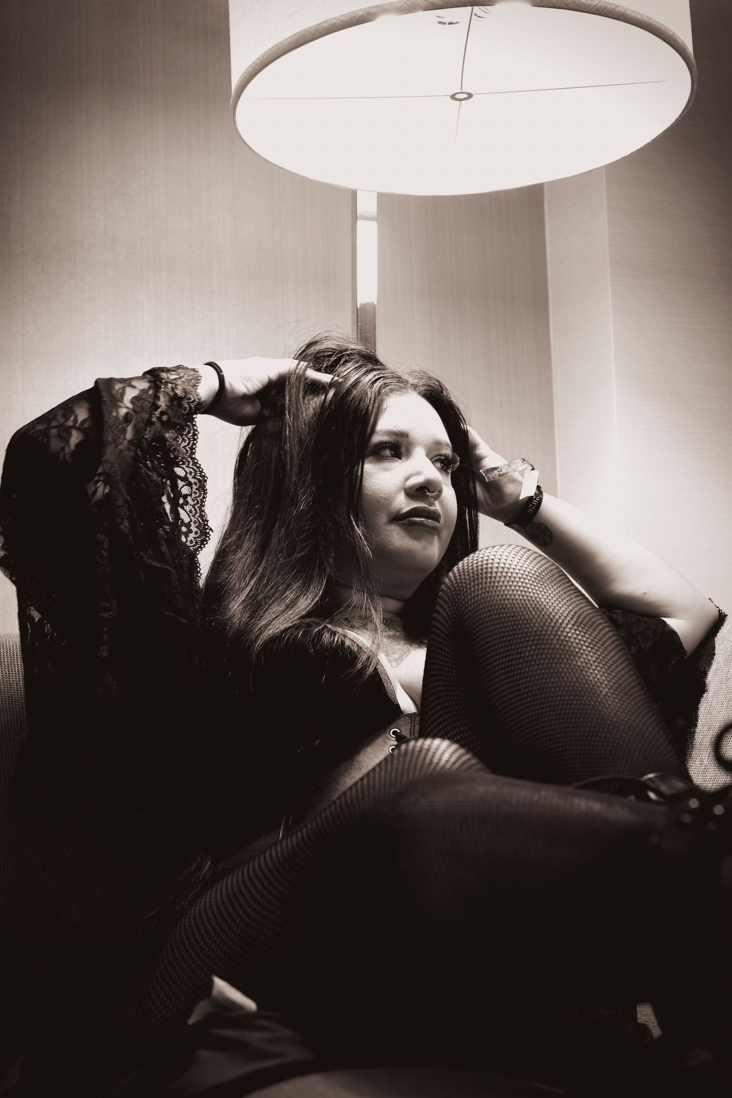 Black and white portrait of a woman in black lace sitting under a lamp in a chair.