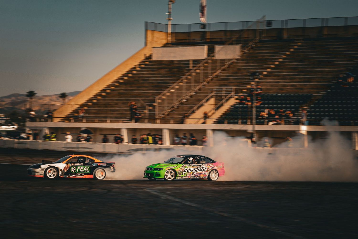 Two drift cars producing massive smoke clouds on a race track with stadium seating behind.