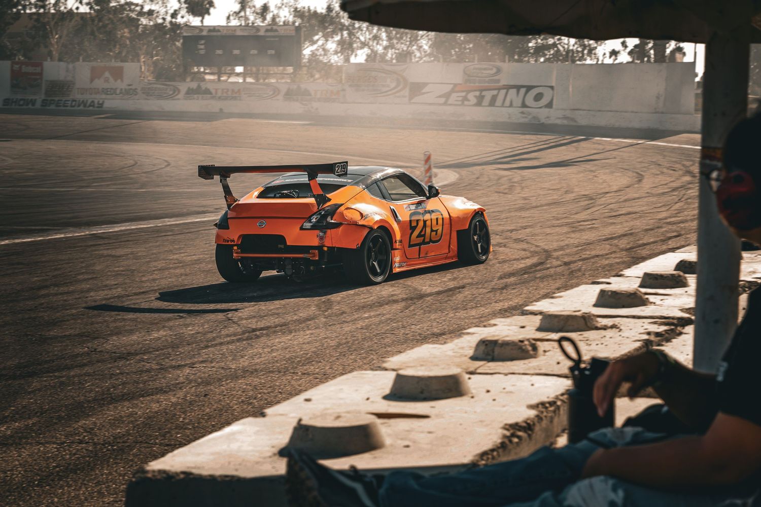 An orange and yellow drift car with number 249 slides across a dusty racing circuit.