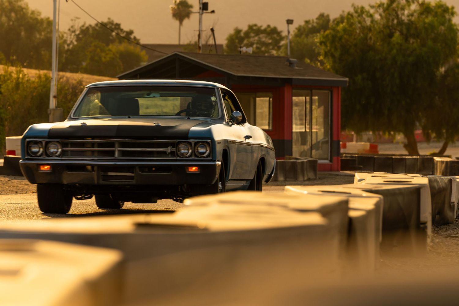 A classic American muscle car parked at golden hour near a vintage drive-in, bathed in warm sunset light.