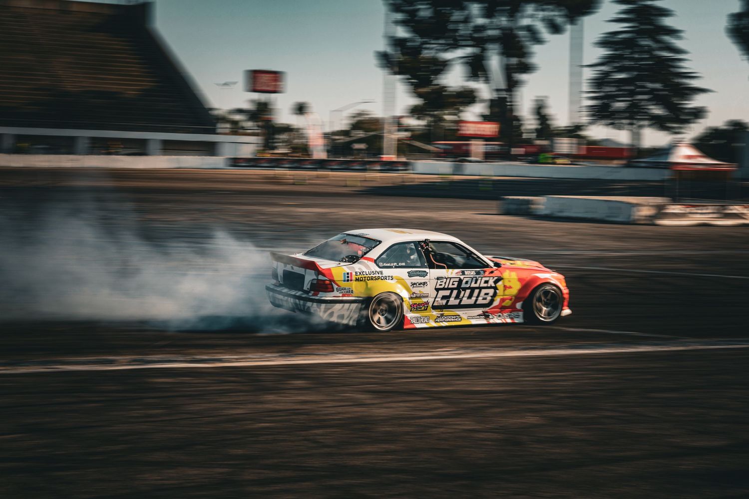 A colorful BMW E36 drift car slides through a corner generating tire smoke on a motorsport track.