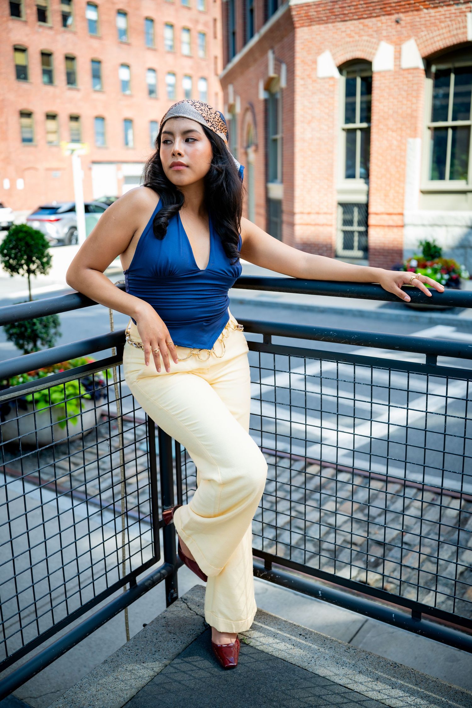 Stylish woman in blue halter and cream wide-leg pants leaning on railing in city setting.