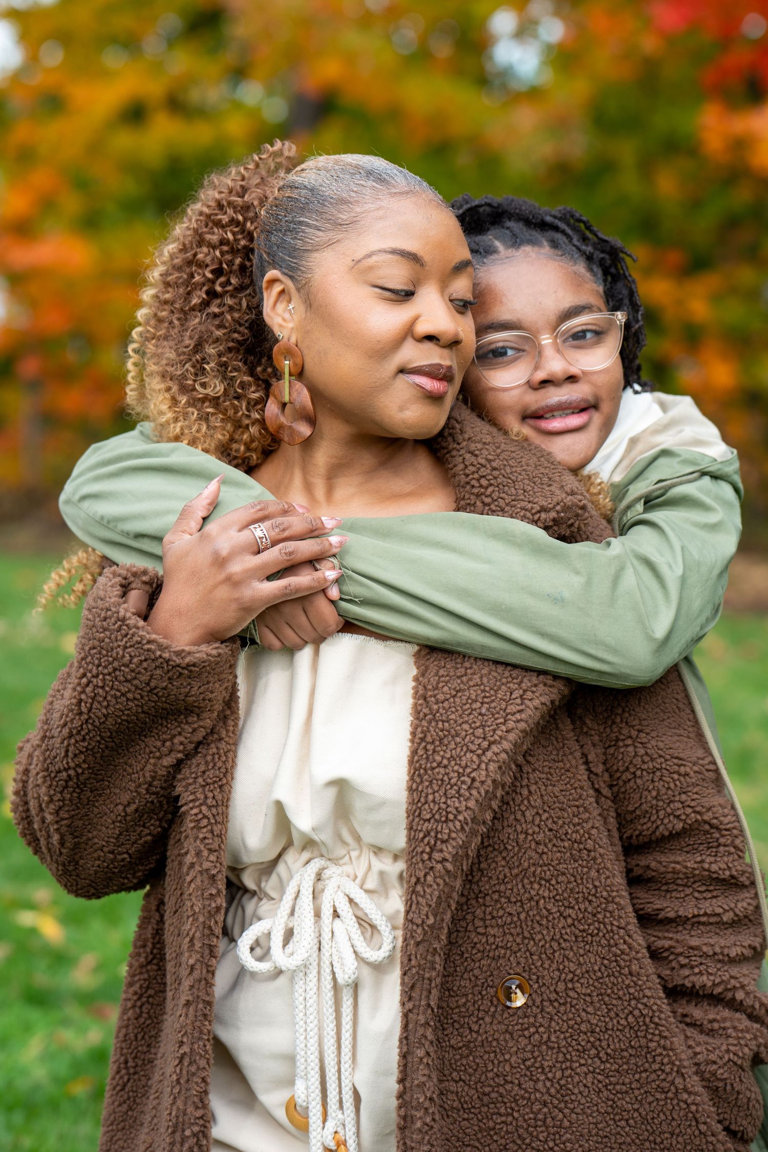 A smiling girl in glasses hugs a woman in a brown sherpa jacket from behind during an outdoor portrait with colorful autumn foliage.