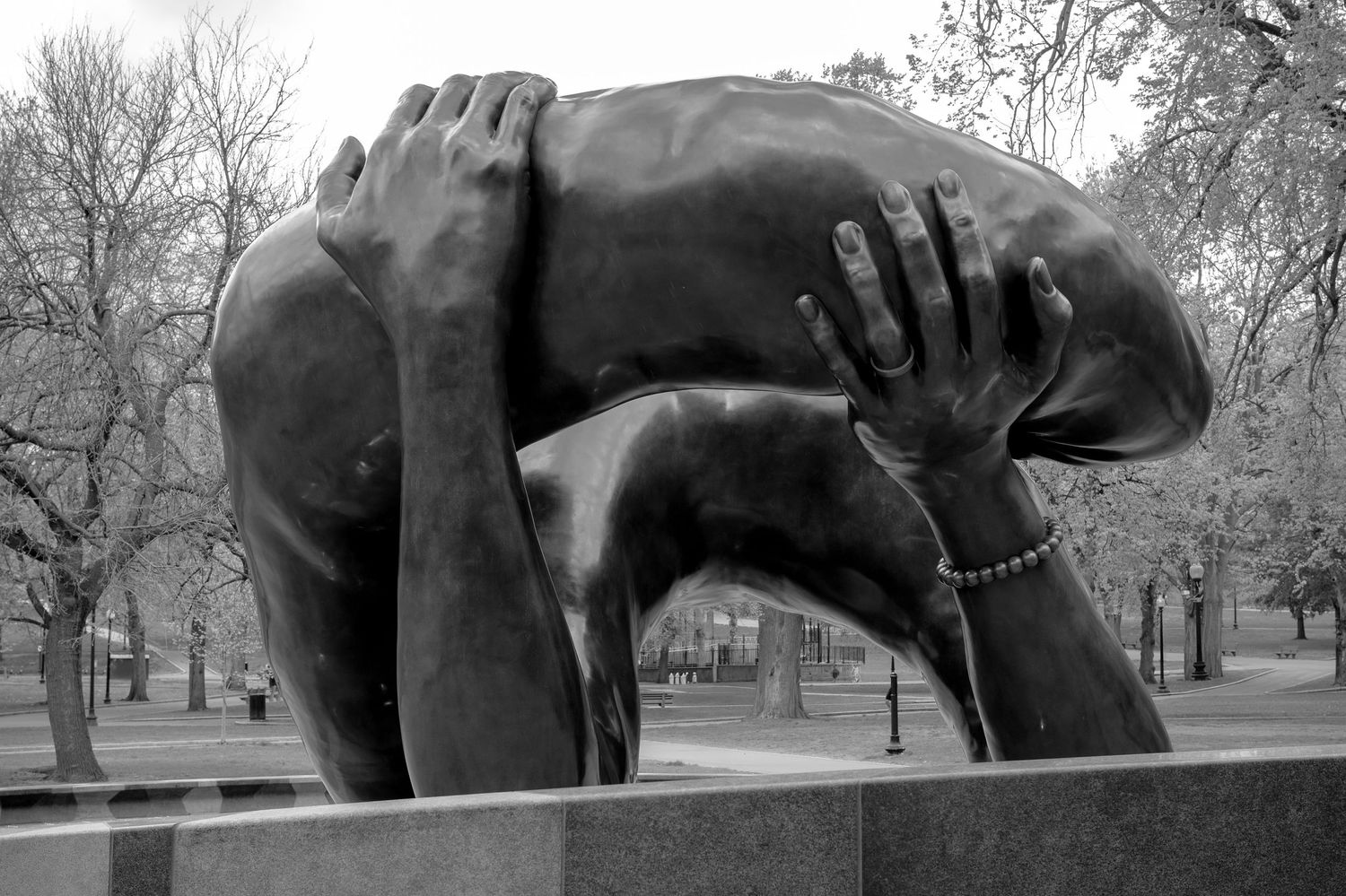 Black and white photo of a bronze sculpture depicting two figures embracing, set in a park with bare trees.