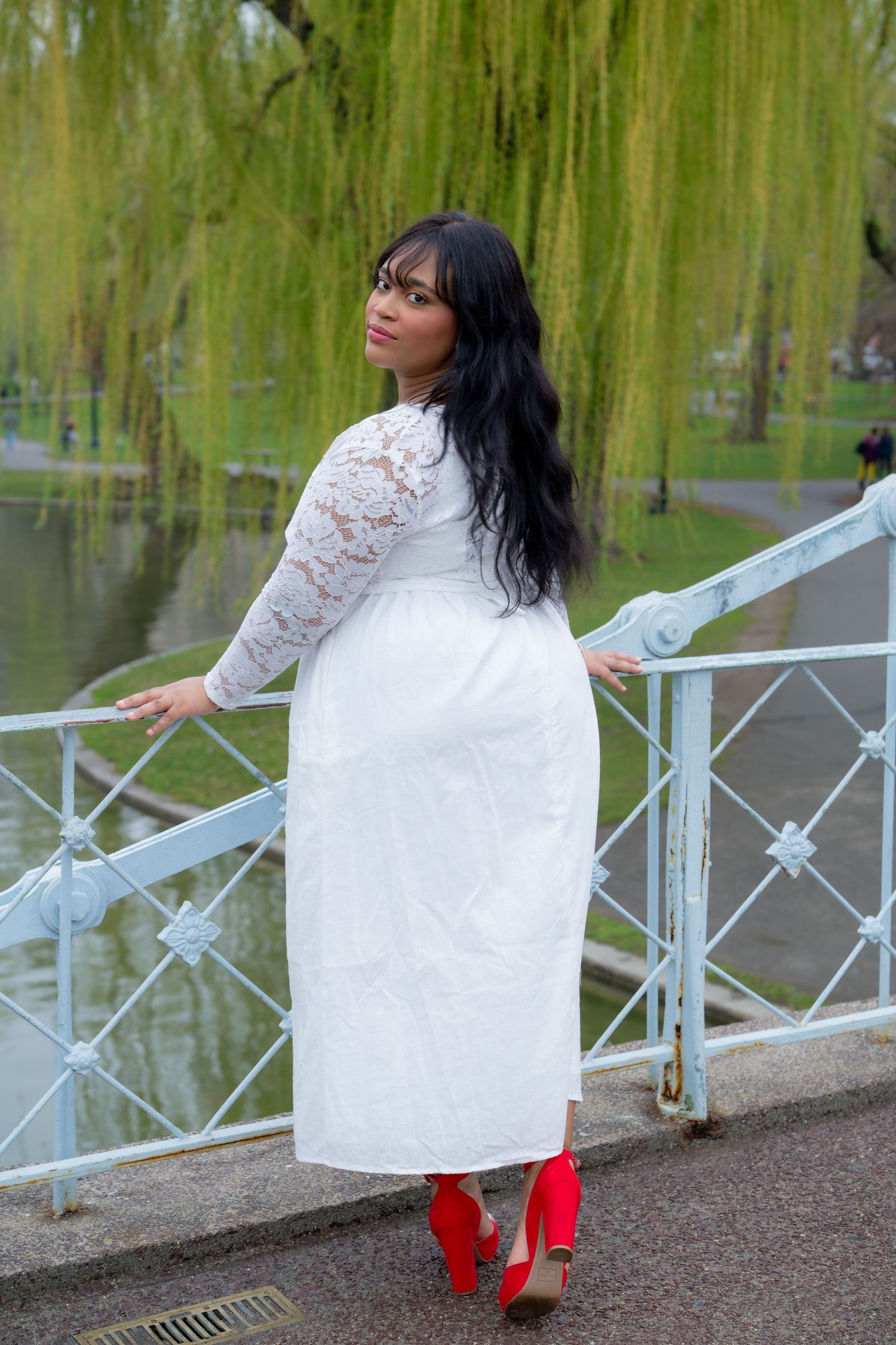 Woman in white lace dress and red heels posing on a bridge by a pond with willow trees.