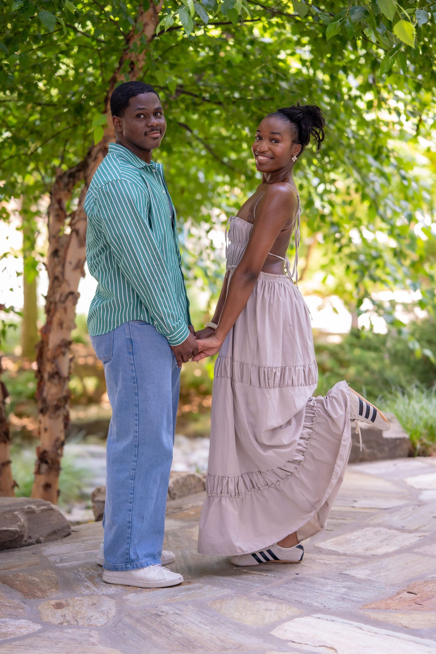 A happy Black couple holds hands and looks back smiling on a stone path surrounded by green trees in summer.