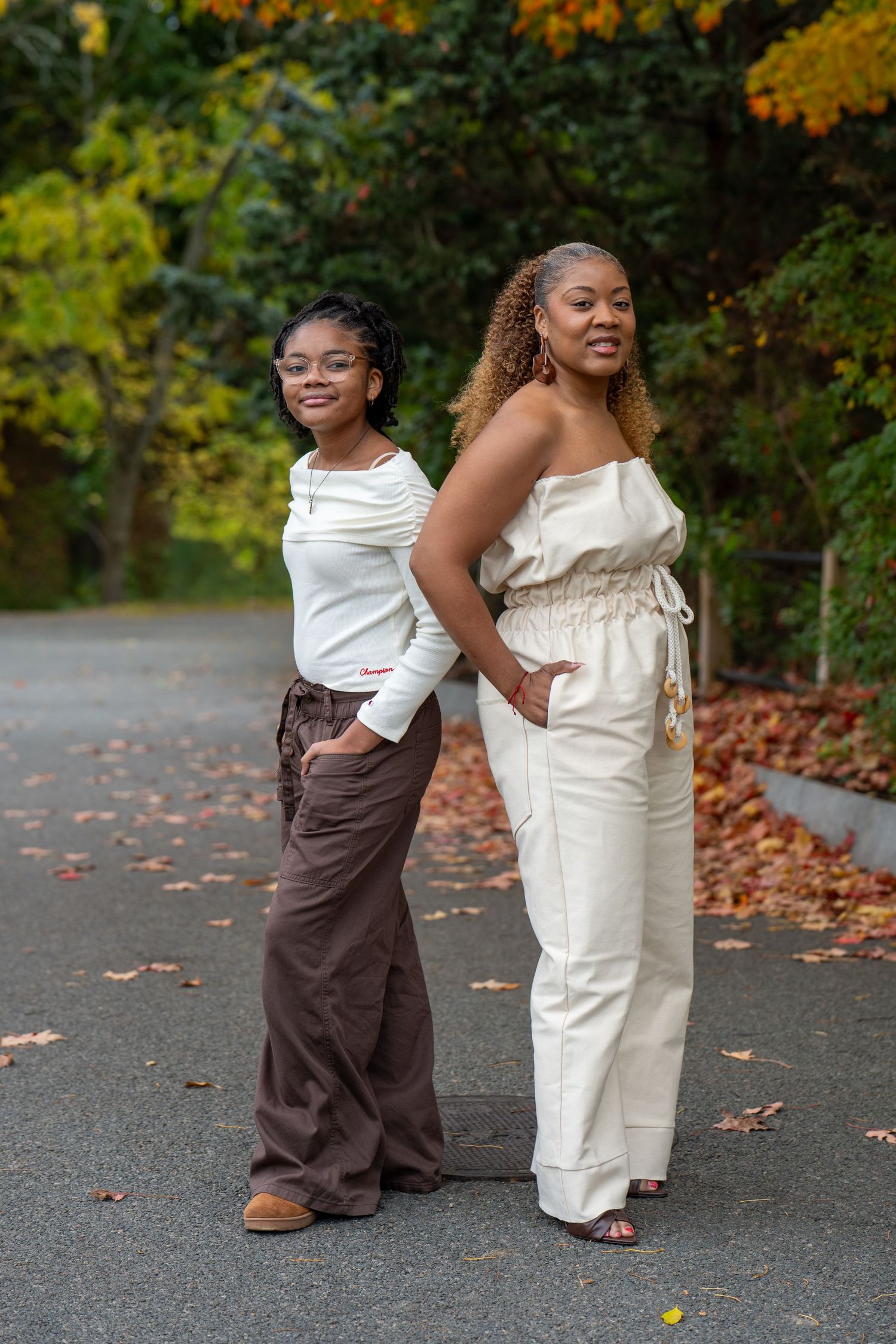 Two women pose back to back on a leaf-covered path in autumn, one in a white top and brown trousers, the other in a cream jumpsuit.