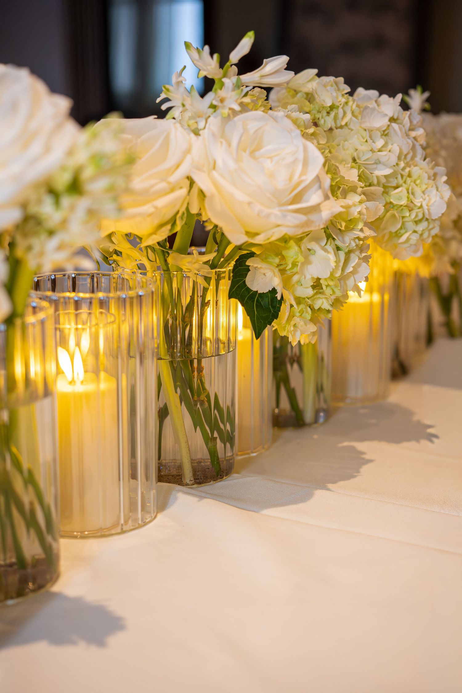 Elegant table centerpiece with white roses, hydrangeas in glass vases and glowing candles on white linen.