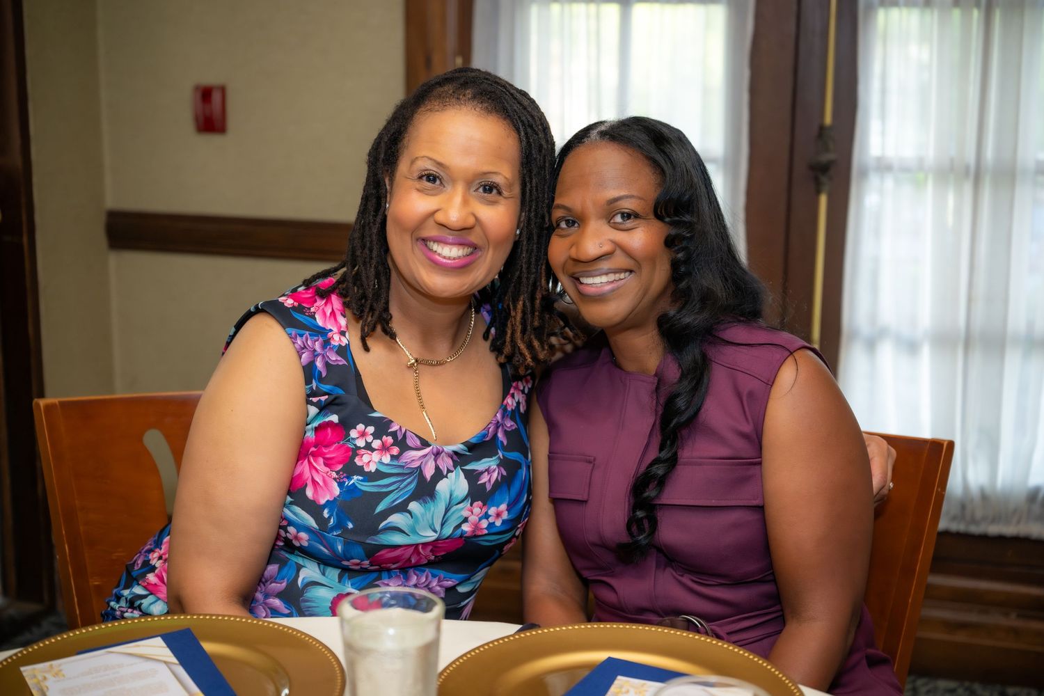 Two smiling women pose together at table during indoor celebration event wearing colorful dresses.