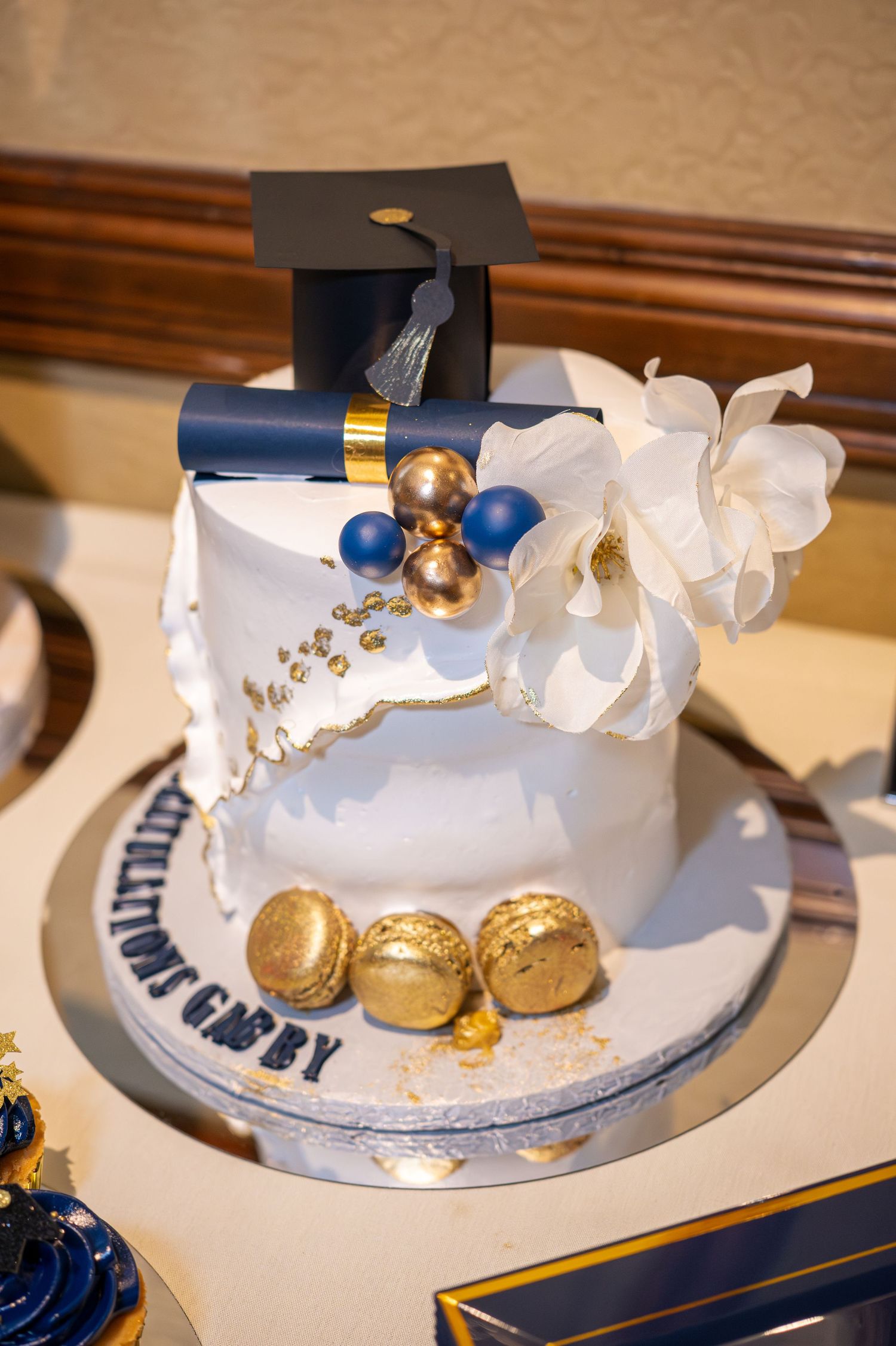 Elegant white graduation cake with gold accents, mortarboard topper, and floral decorations.
