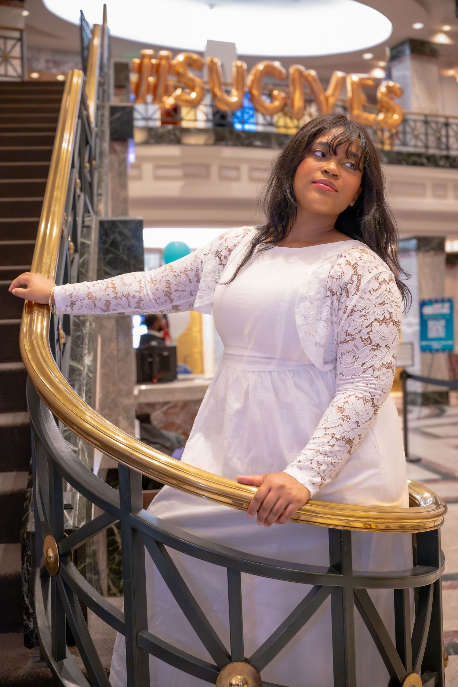 Young woman in white lace dress poses gracefully on golden staircase at Suffolk University event.
