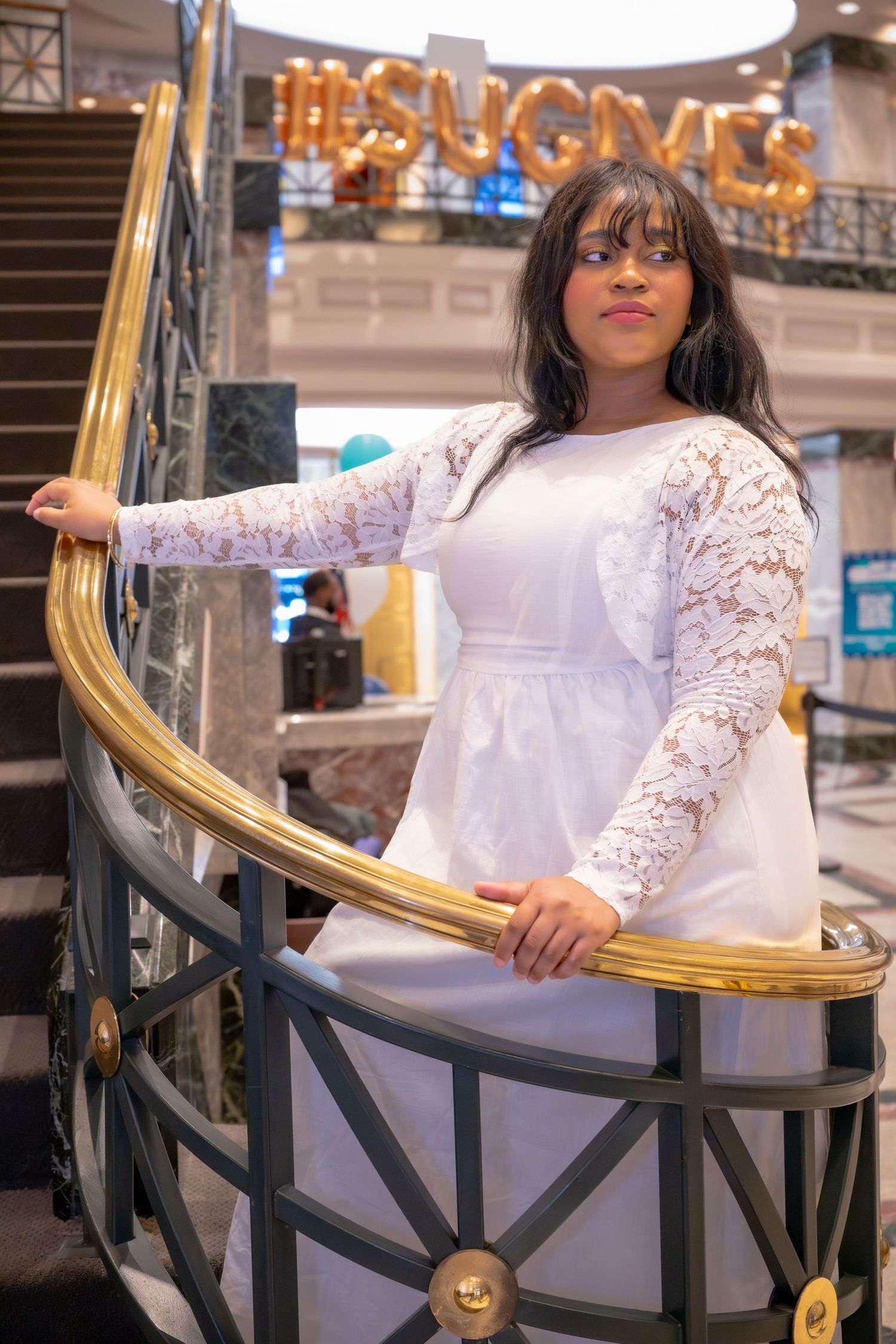 A young Black woman in a white lace dress stands on a curved staircase with a gold railing at an event venue.