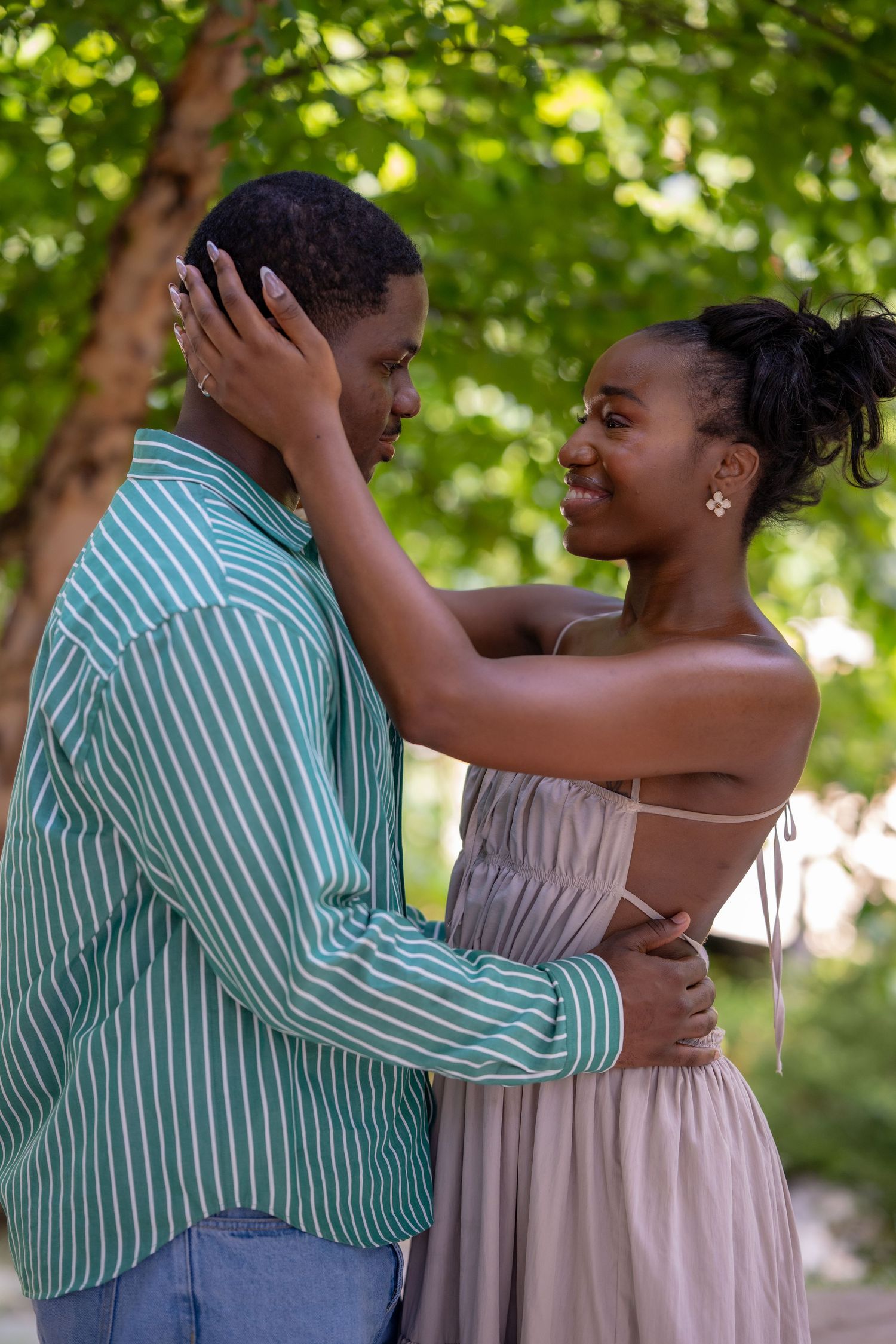 A smiling Black couple embraces outdoors, the woman in a pink maxi dress holding the mans face near lush green trees.
