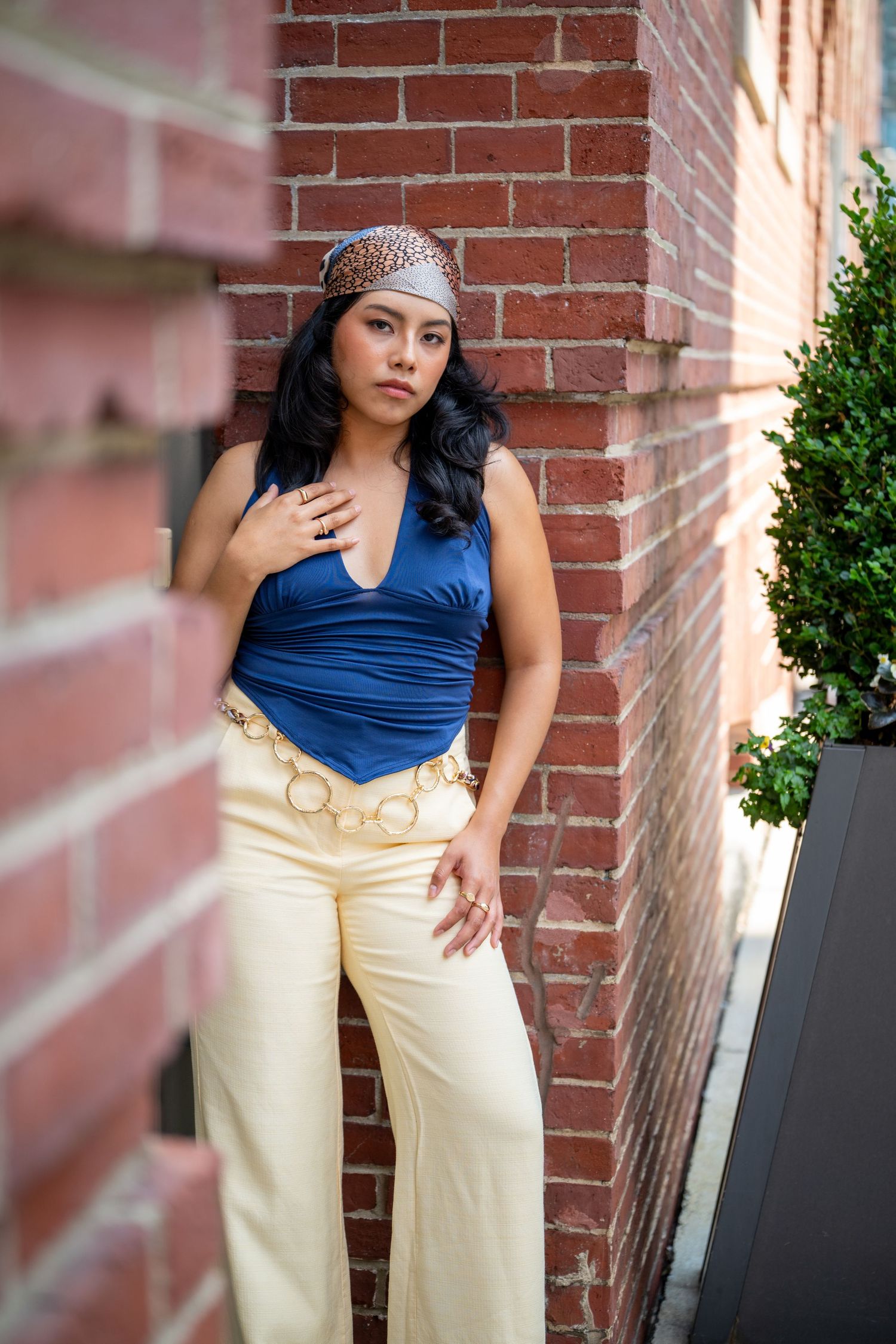 Stylish woman in blue halter top and cream wide-leg pants poses by brick wall.