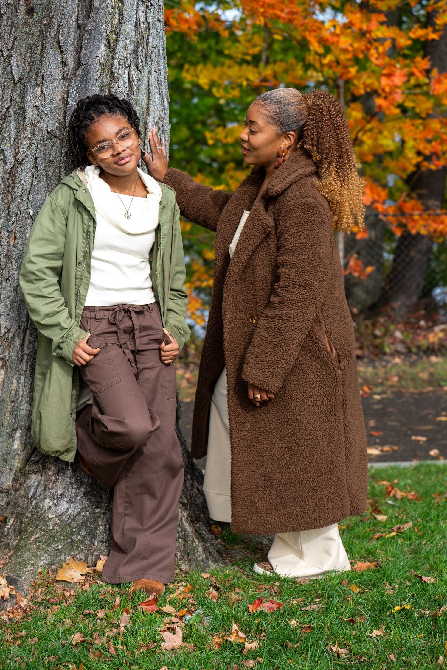 Two women in earth-toned coats lean against a large tree trunk on a grassy lawn, with vibrant orange autumn leaves in the background.