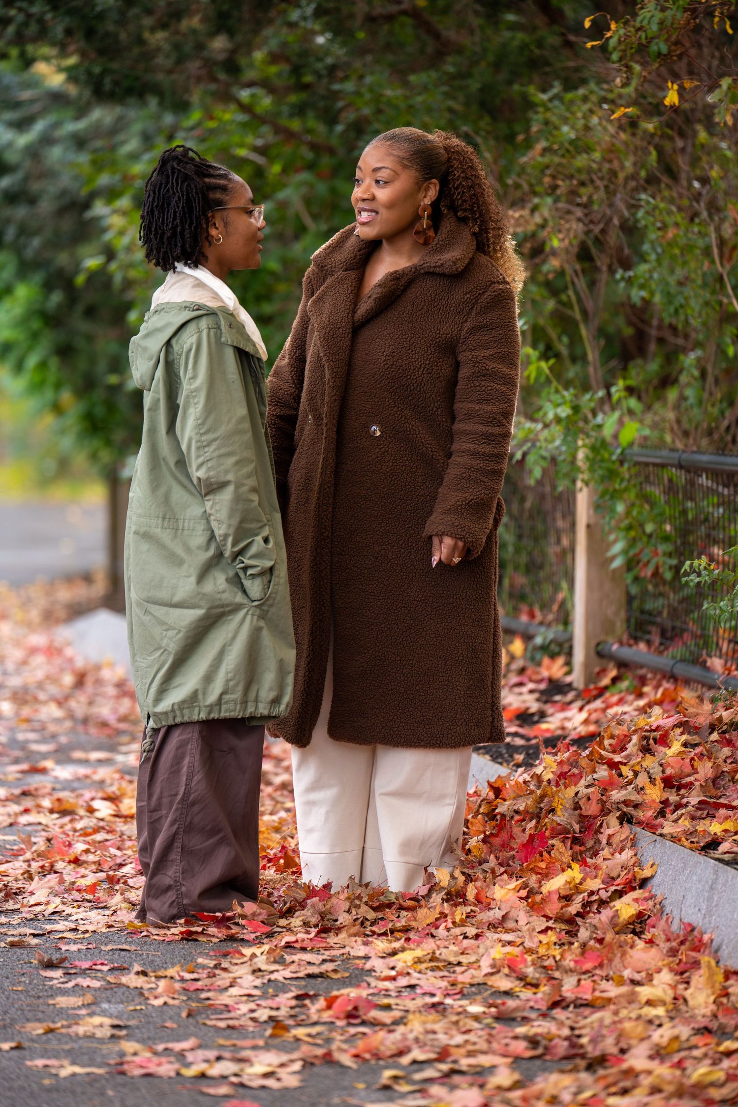 Two women in cozy autumn coats stand face to face and talk on a leaf-covered path lined with colorful fall foliage.