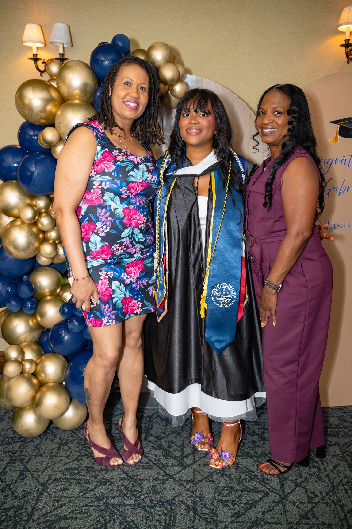 Three women pose together at a graduation celebration, with the graduate in cap and gown flanked by two smiling guests.