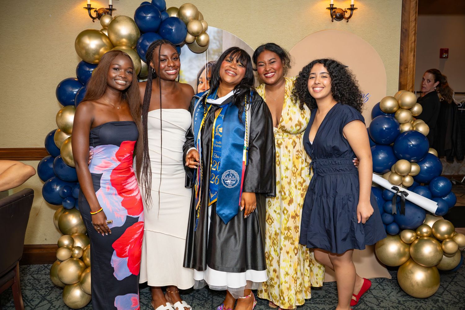 Graduate in cap and gown celebrates with four friends at a graduation party with festive balloons.