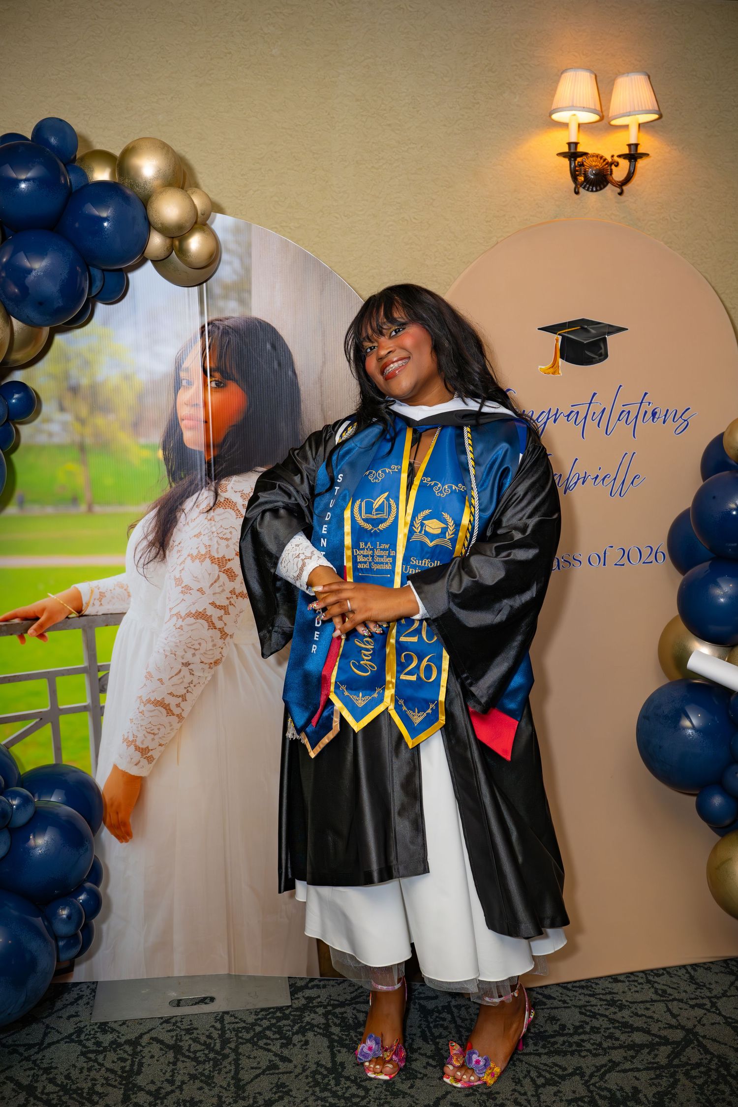 Graduate in blue and gold regalia poses beside large photo cutout at Suffolk University Class of 2026 party.