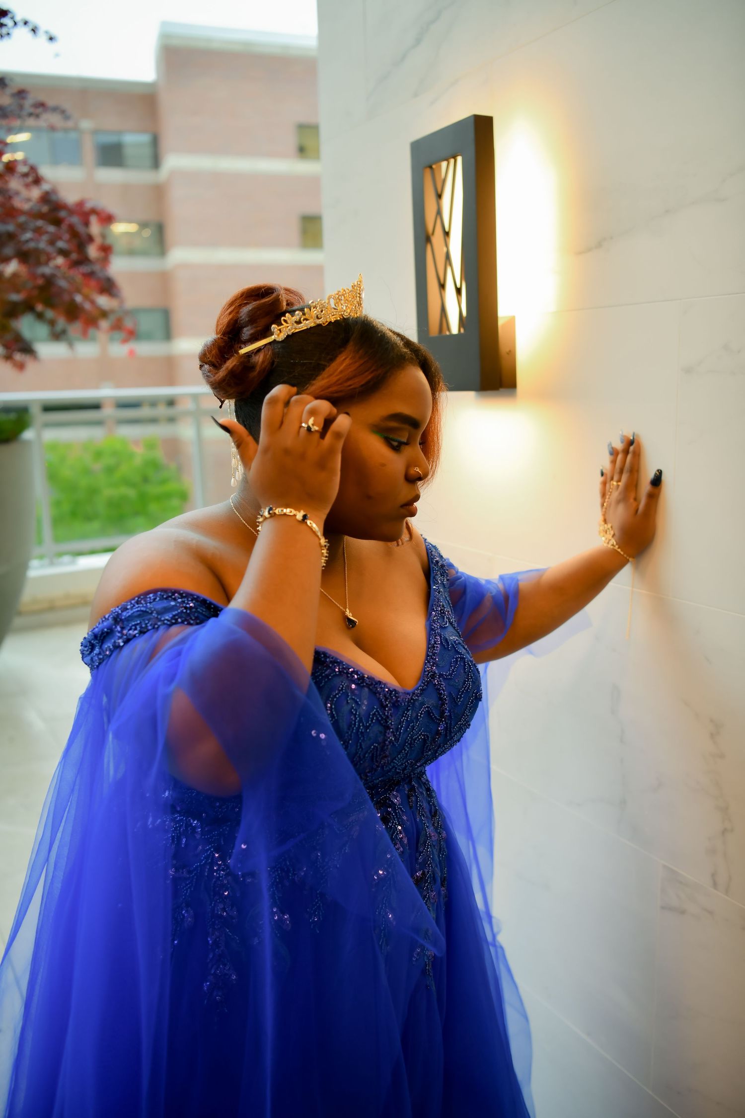 A woman in a royal blue off-shoulder gown and crown leans gracefully against a wall.