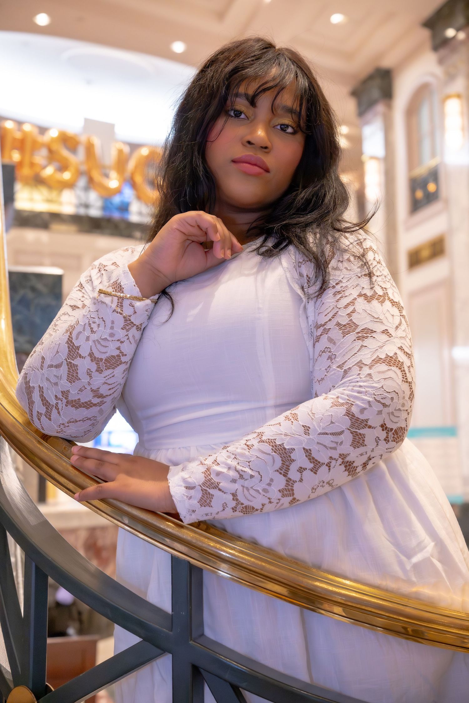 A young Black woman in a white lace dress poses elegantly on a curved gold staircase railing indoors.